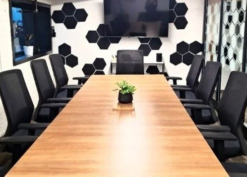 Conference room with a wooden table, black mesh chairs, a potted plant, a wall-mounted TV, and decorative honeycomb-patterned walls.