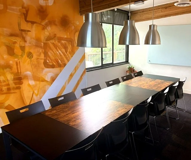 A modern conference room with a rectangular wooden table, six white chairs, a large window with a gray shade, a wall-mounted TV, three gray pendant lights, and an orange and yellow mural on one wall.