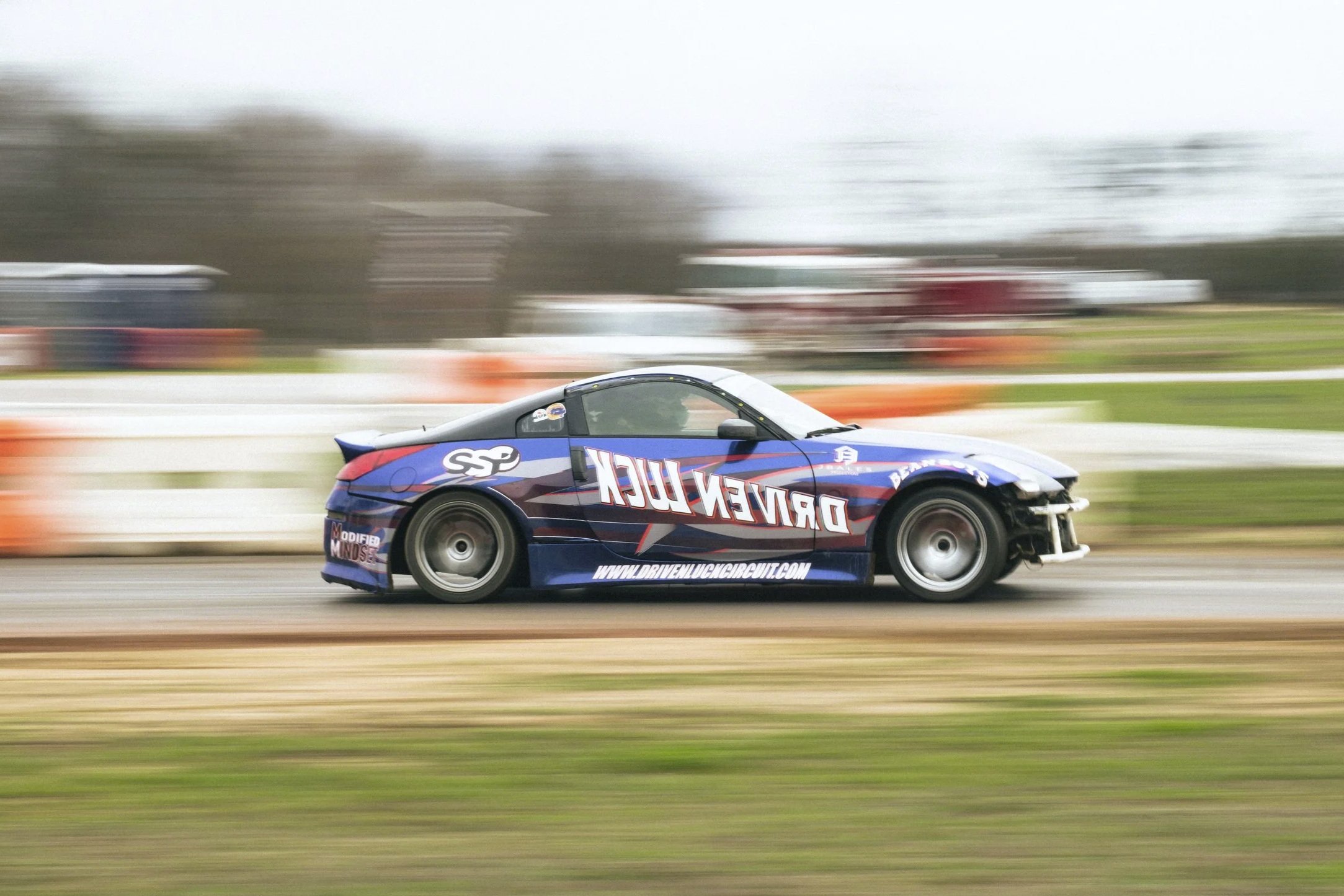A race car speeding on a track with motion blur, featuring a blue and red design and various sponsor logos.