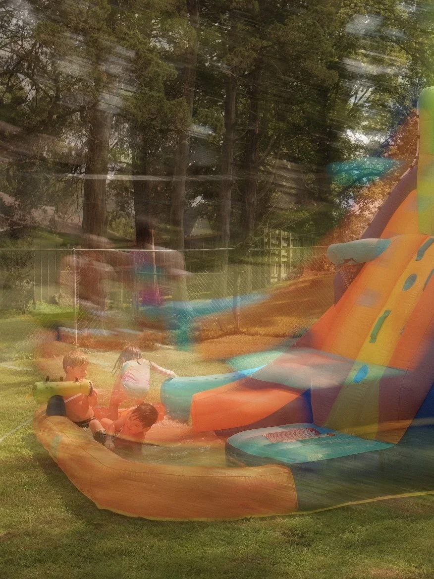 Children playing on an inflatable water slide in a backyard with trees and a fence.