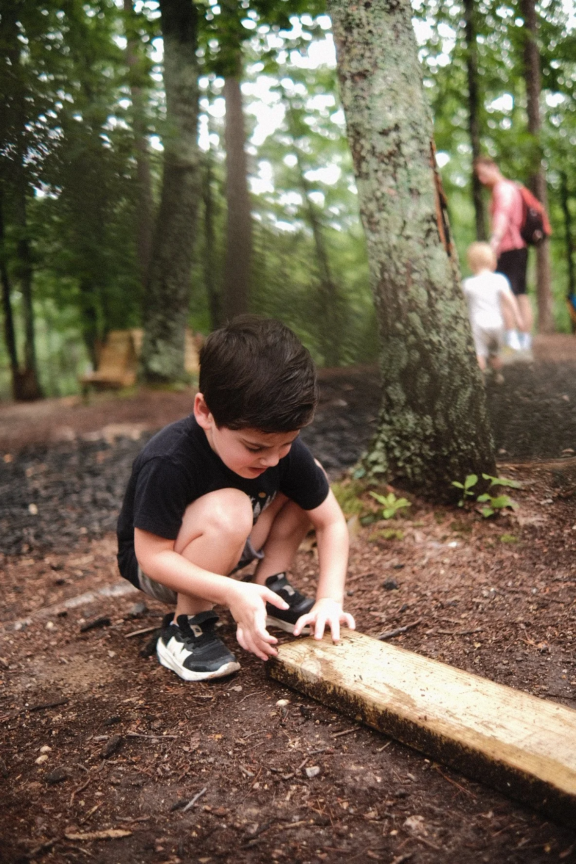 Young boy in black shirt and shorts crouches on dirt ground in a forest, inspecting or working on a piece of wood on the ground. Behind him, a woman and a young child are walking along a trail in the woods.