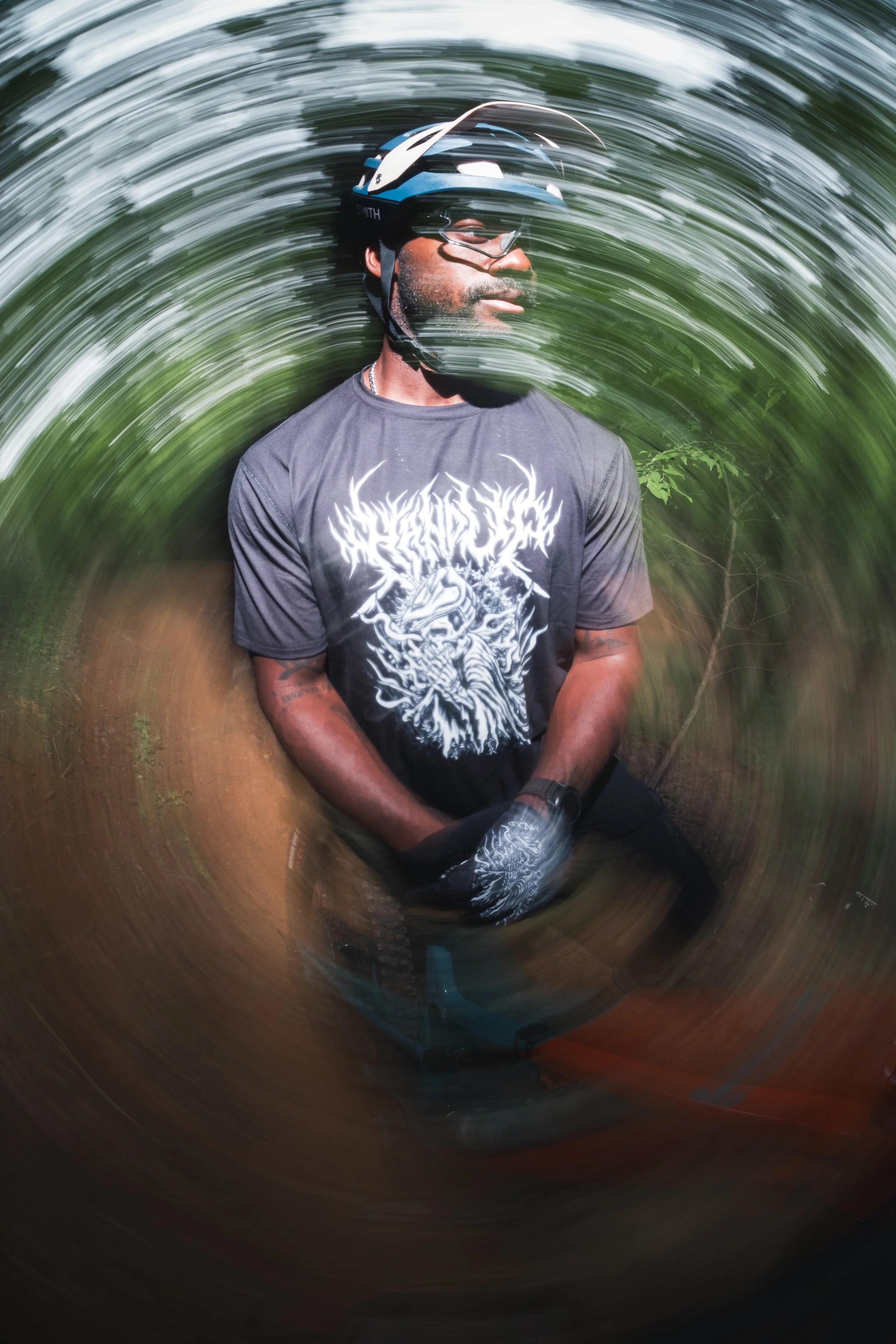 A man wearing a helmet and sunglasses, sitting outdoors in a forested area, with a swirling motion effect around him.