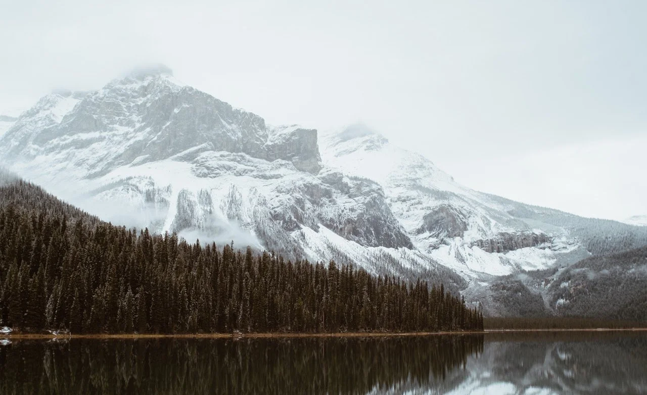 Snow-covered mountain behind a forest of evergreen trees reflected in a lake.