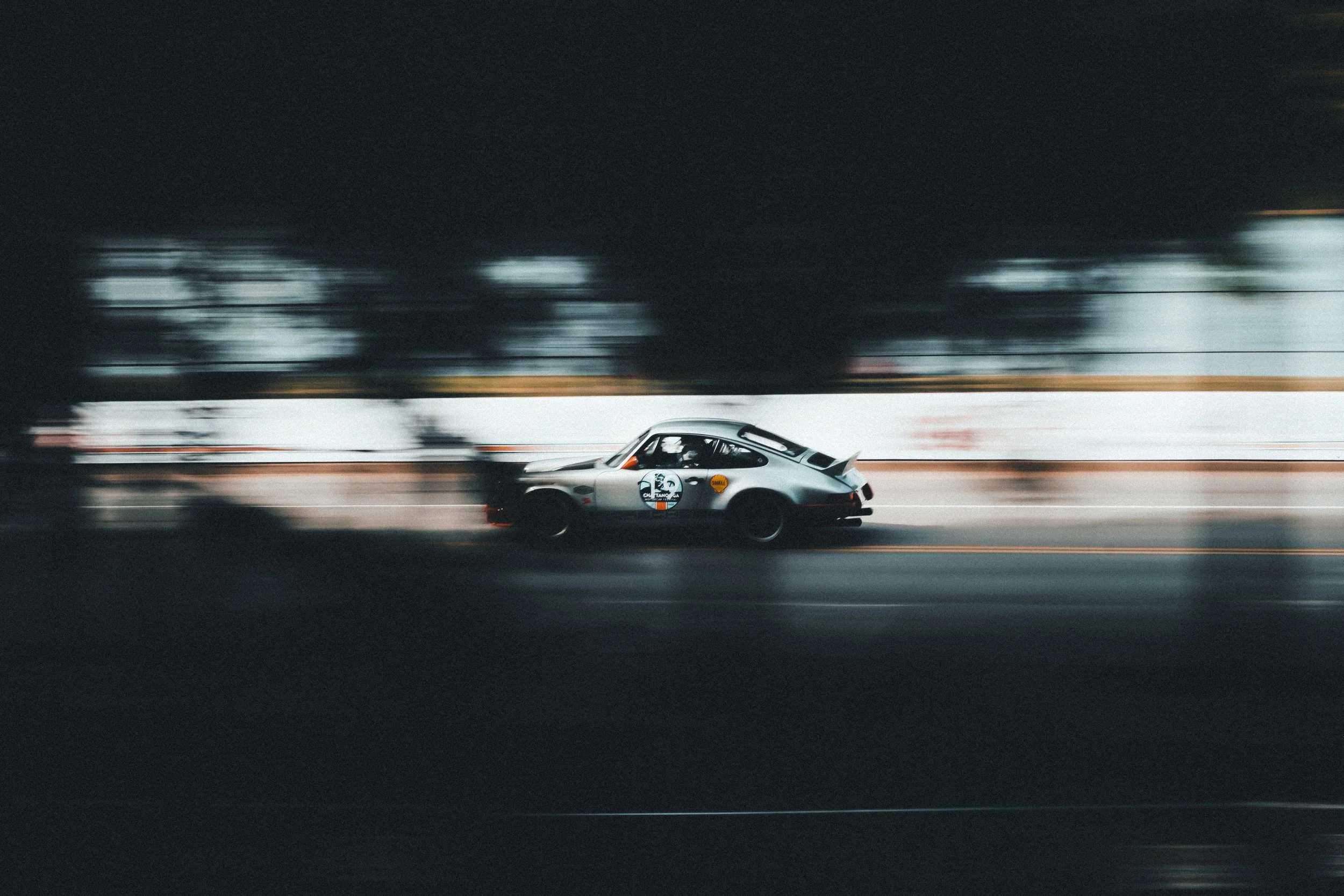 A silver race car with a racing number and logo on the side, speeding along a track, with motion blur in the background.
