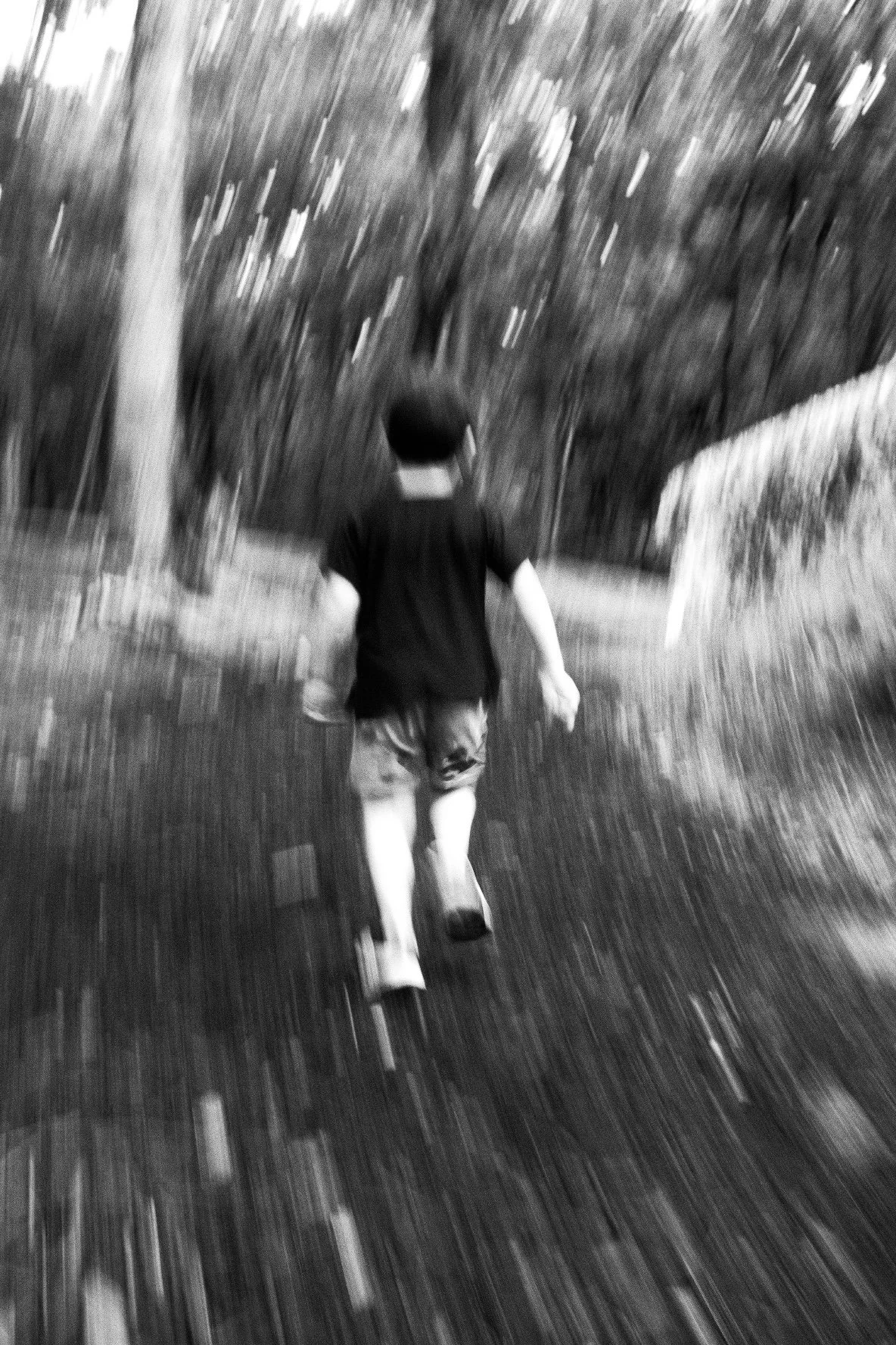 A black and white photo of a person walking along a dirt path through a wooded area, viewed from behind, with a slightly blurred motion effect.