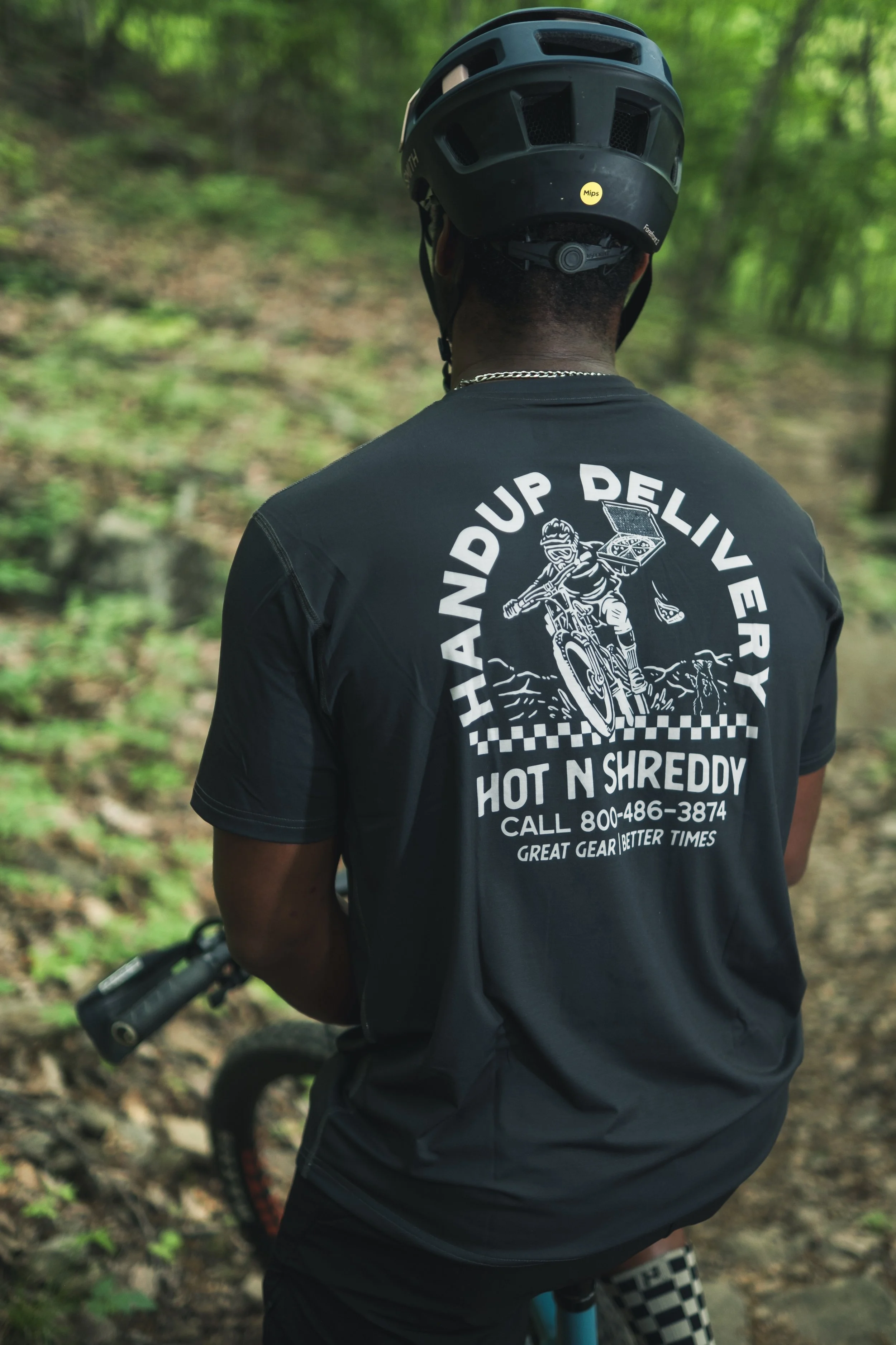 A person wearing a black helmet and a black t-shirt with a graphic and text on the back, riding a mountain bike through a wooded trail.