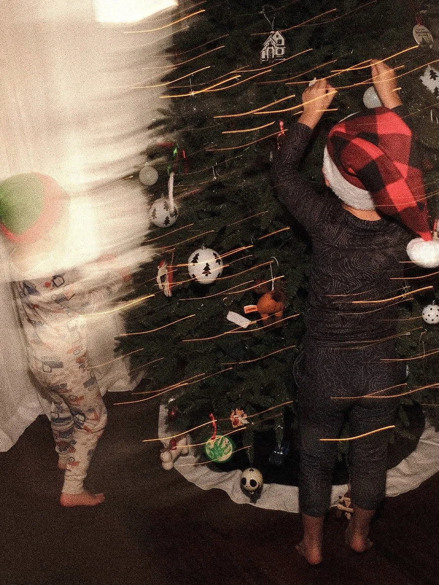 Two children decorating a Christmas tree indoors, one wearing pajamas and the other wearing a Santa hat and pajamas, with festive ornaments on the tree.