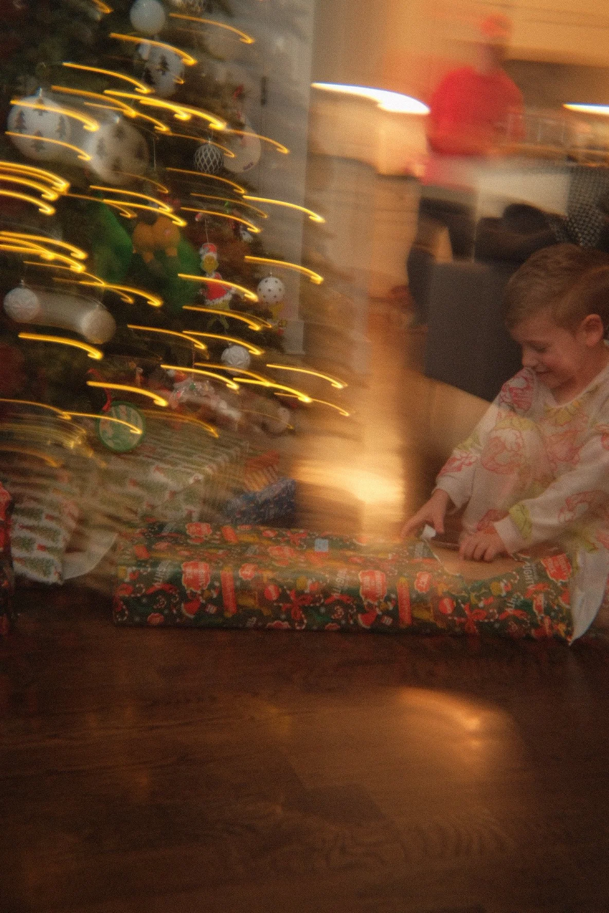 A boy opening a Christmas gift with a Christmas tree in the background decorated with lights and ornaments.
