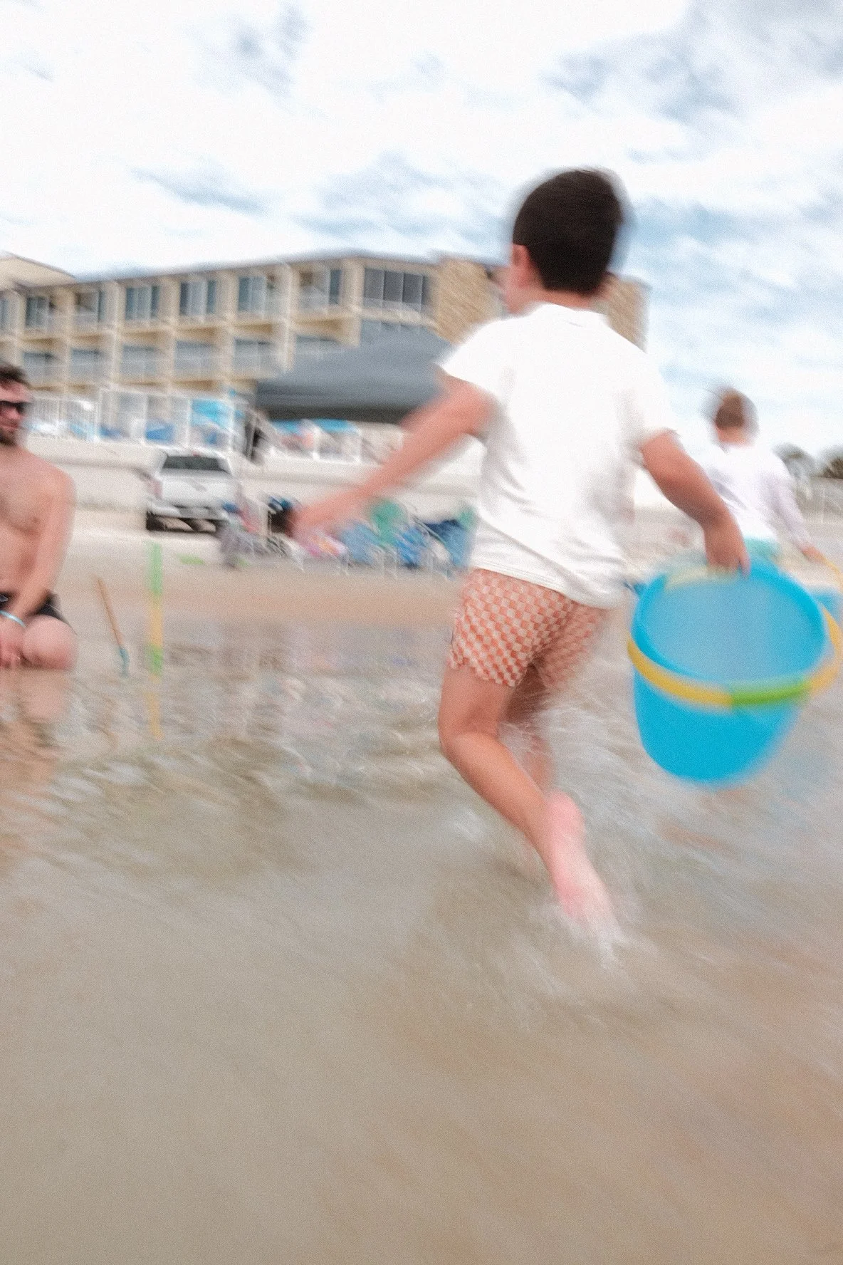 Child running through shallow water on the beach holding a blue and yellow bucket, with adults and beach resort buildings in the background.