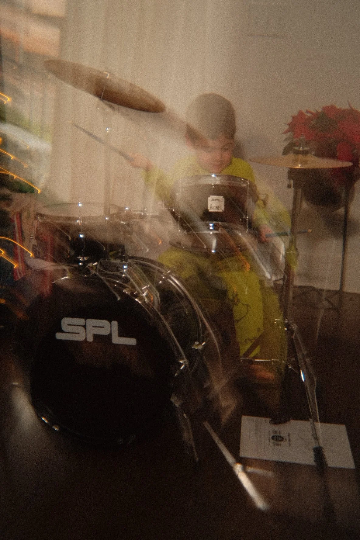A young boy dressed in a yellow outfit sitting at a drum set, with Christmas decorations nearby.
