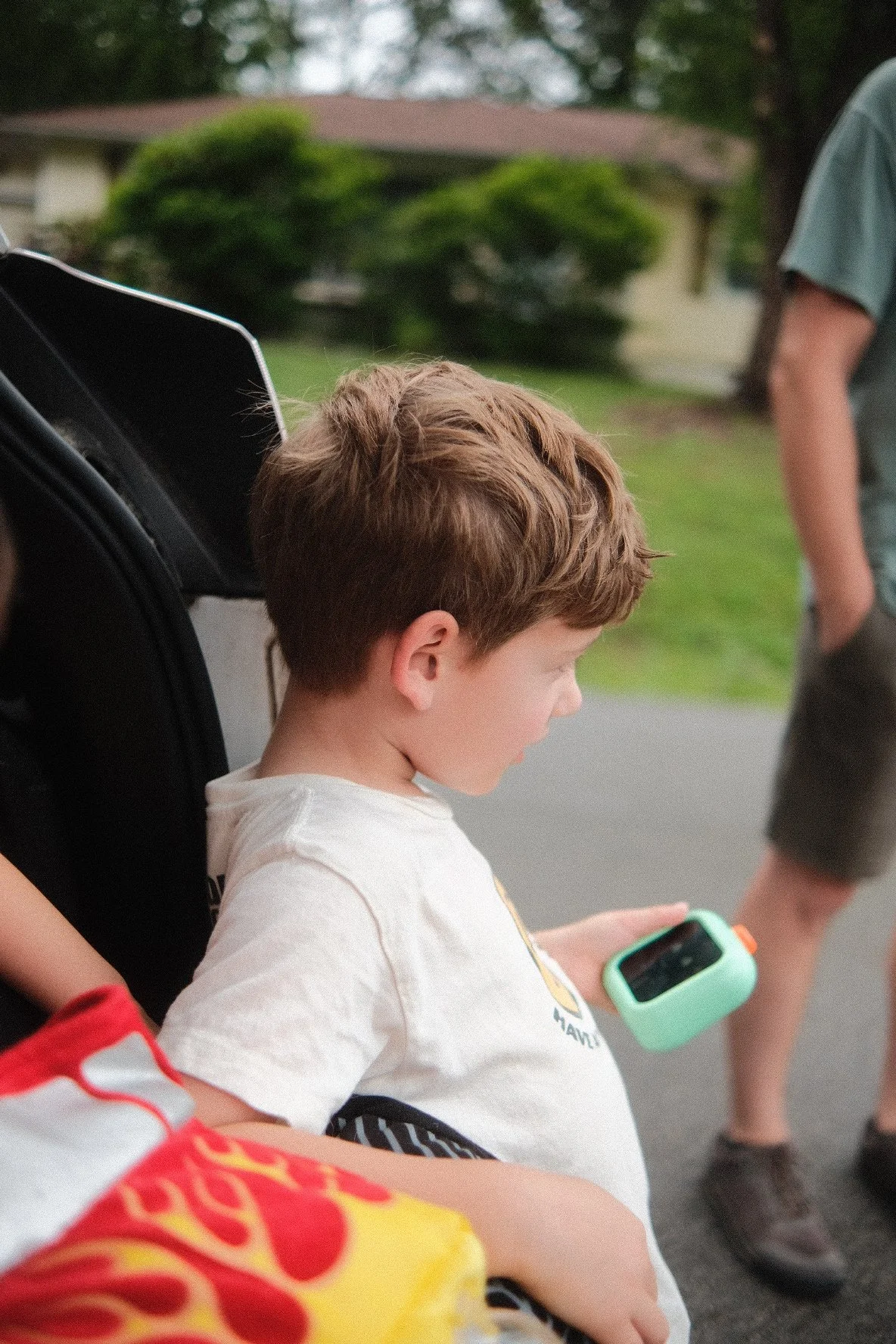 A young boy with brown hair, wearing a white shirt, is sitting in a wheelchair and playing with a handheld gaming device outdoors.