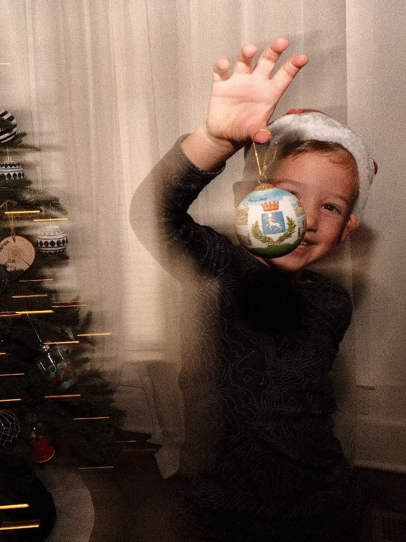 A boy wearing a Santa hat holding a Christmas ornament in front of a decorated Christmas tree.