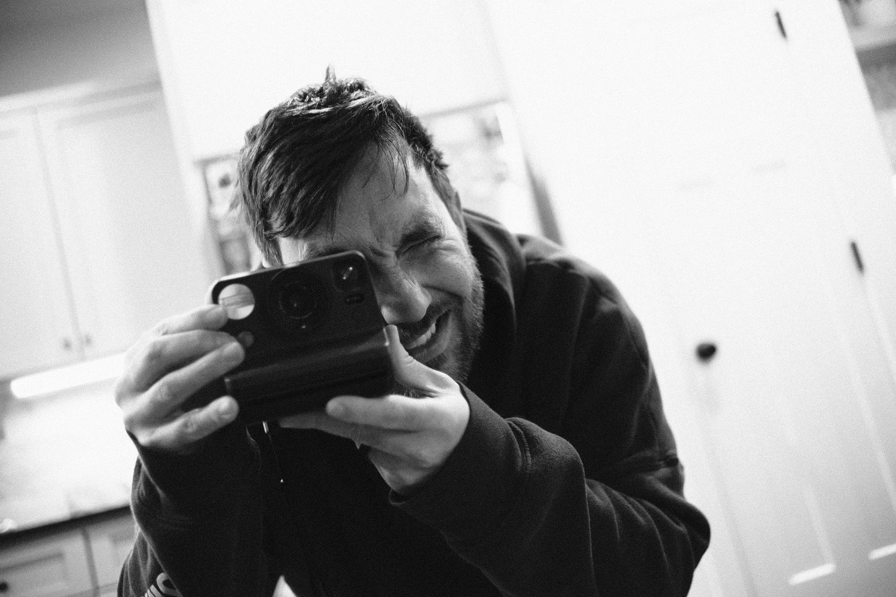 A man taking a selfie using a smartphone, smiling and squinting, indoors with cabinets in the background.