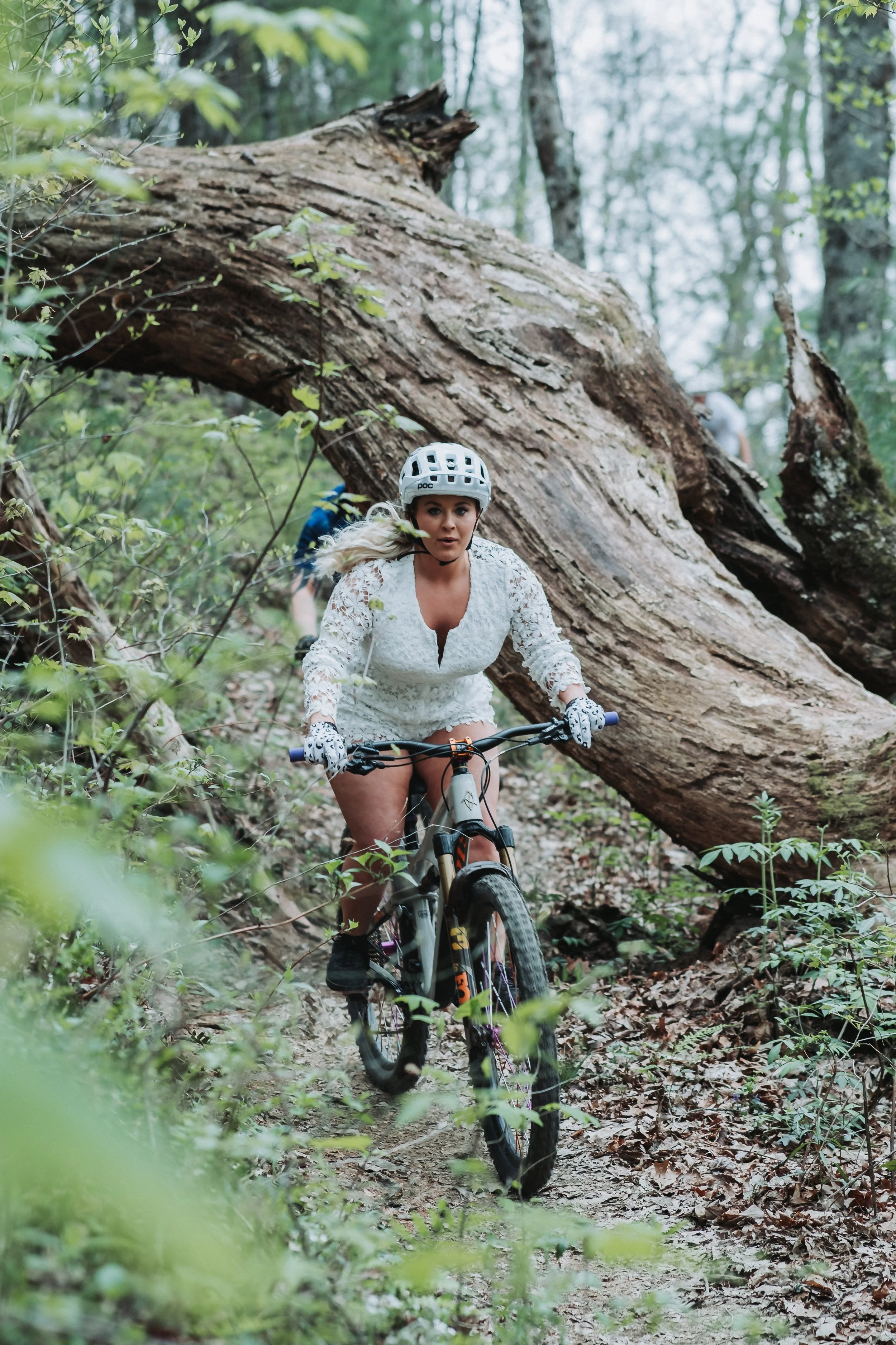 A woman mountain biking through a forest, wearing a white lace top, helmet, and gloves, with fallen trees and green plants around her.