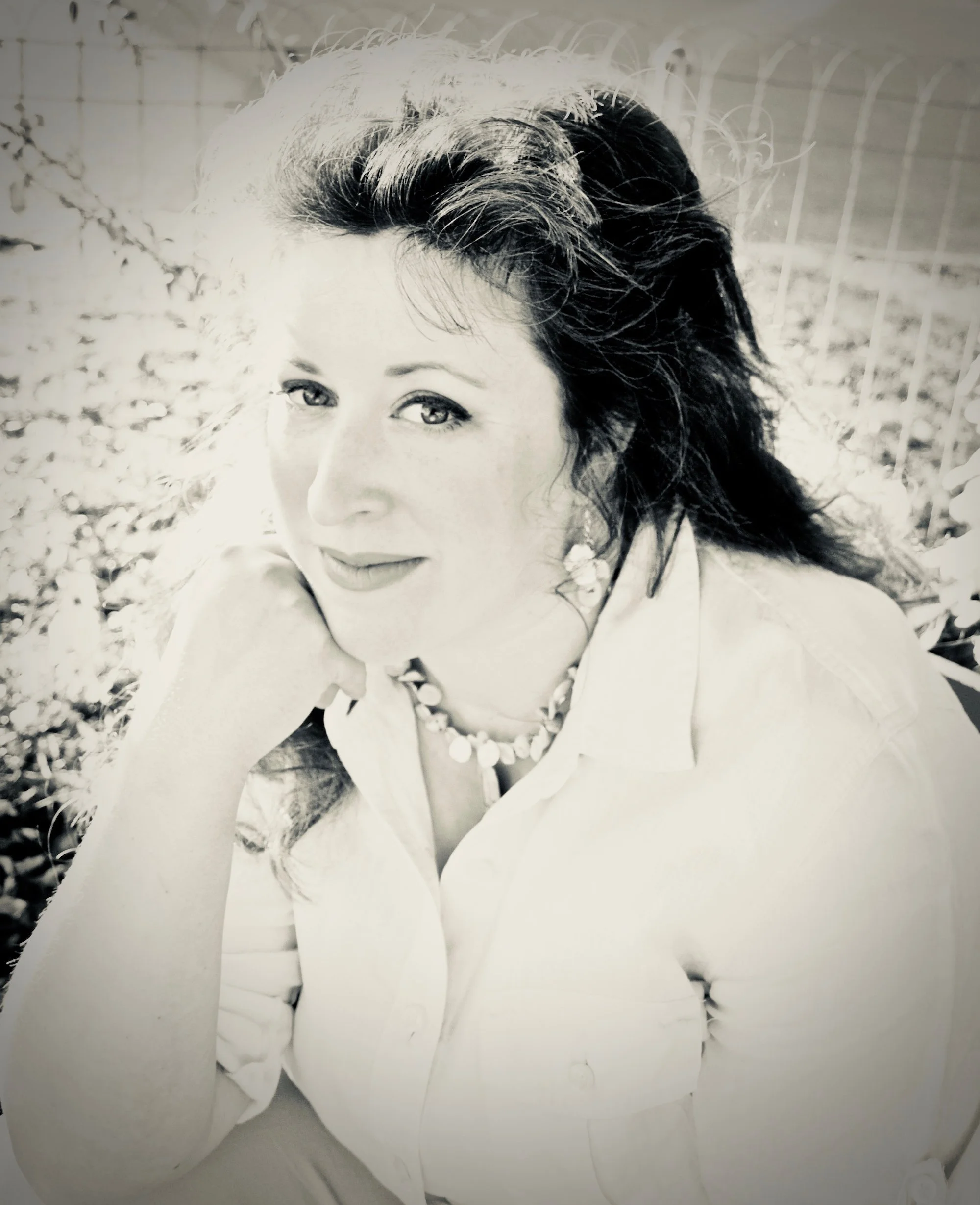 A black-and-white portrait of a woman with dark, wavy hair, wearing a pearl necklace and a light-colored shirt, resting her chin on her hand outdoors.