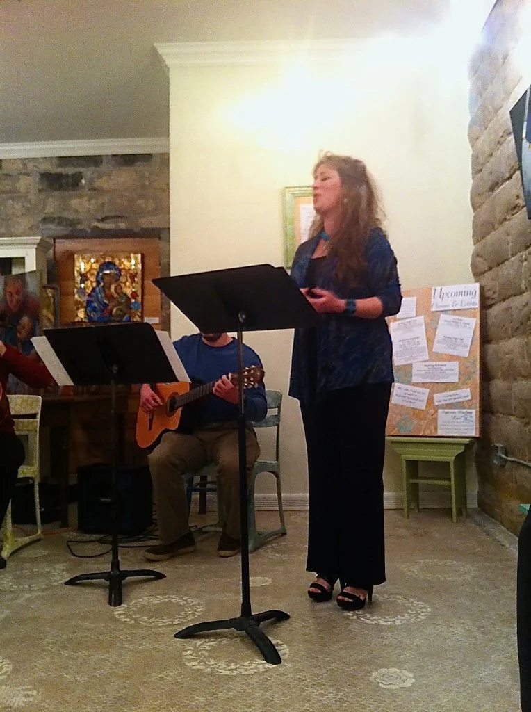 A woman standing and singing in front of a music stand, with a man playing guitar behind her, during a musical performance indoors. Dr. Ada Hunter, Soprano.