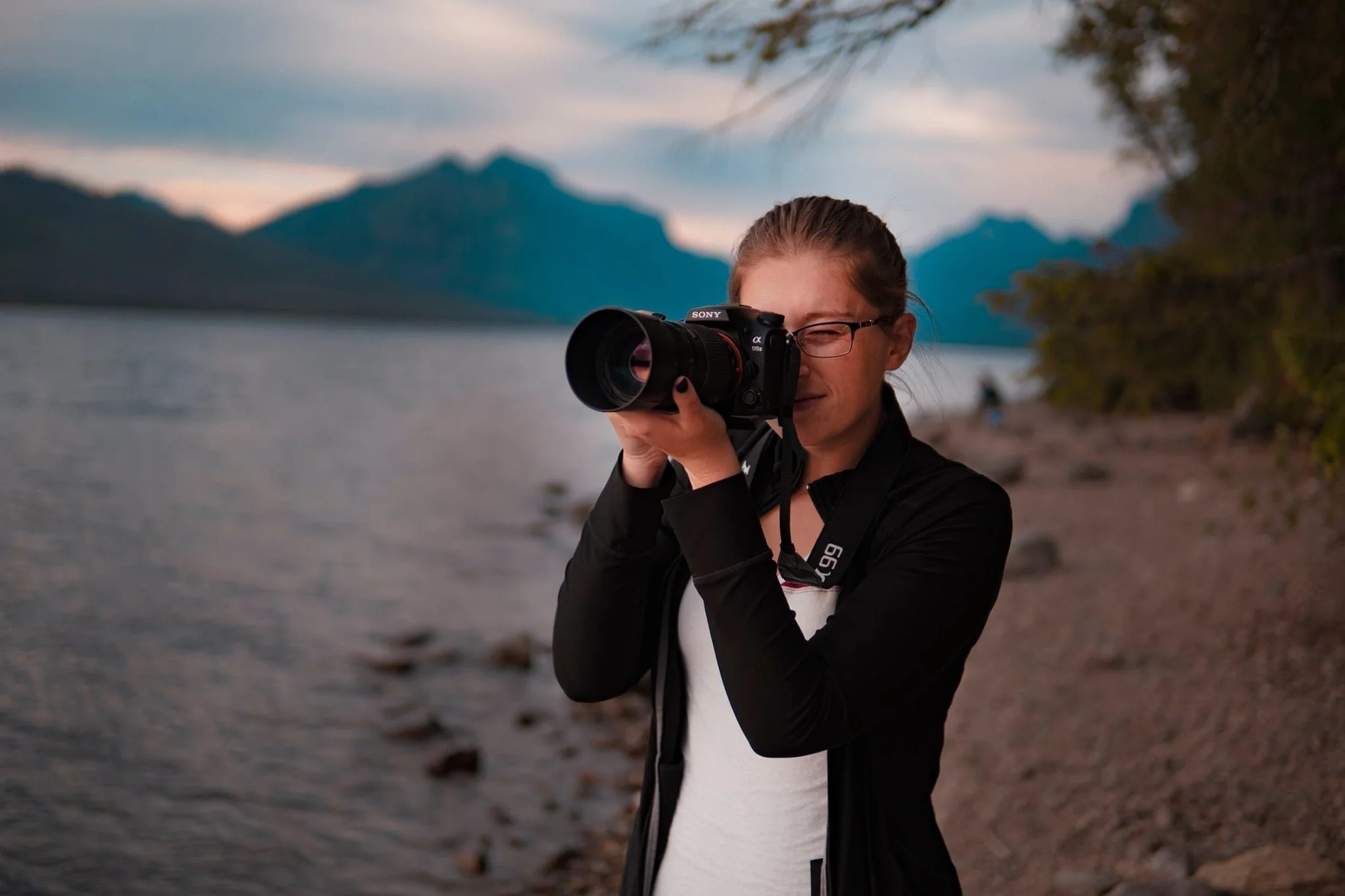 Caitlyn Hymer taking a photo with a camera by a lake, mountains in the background.