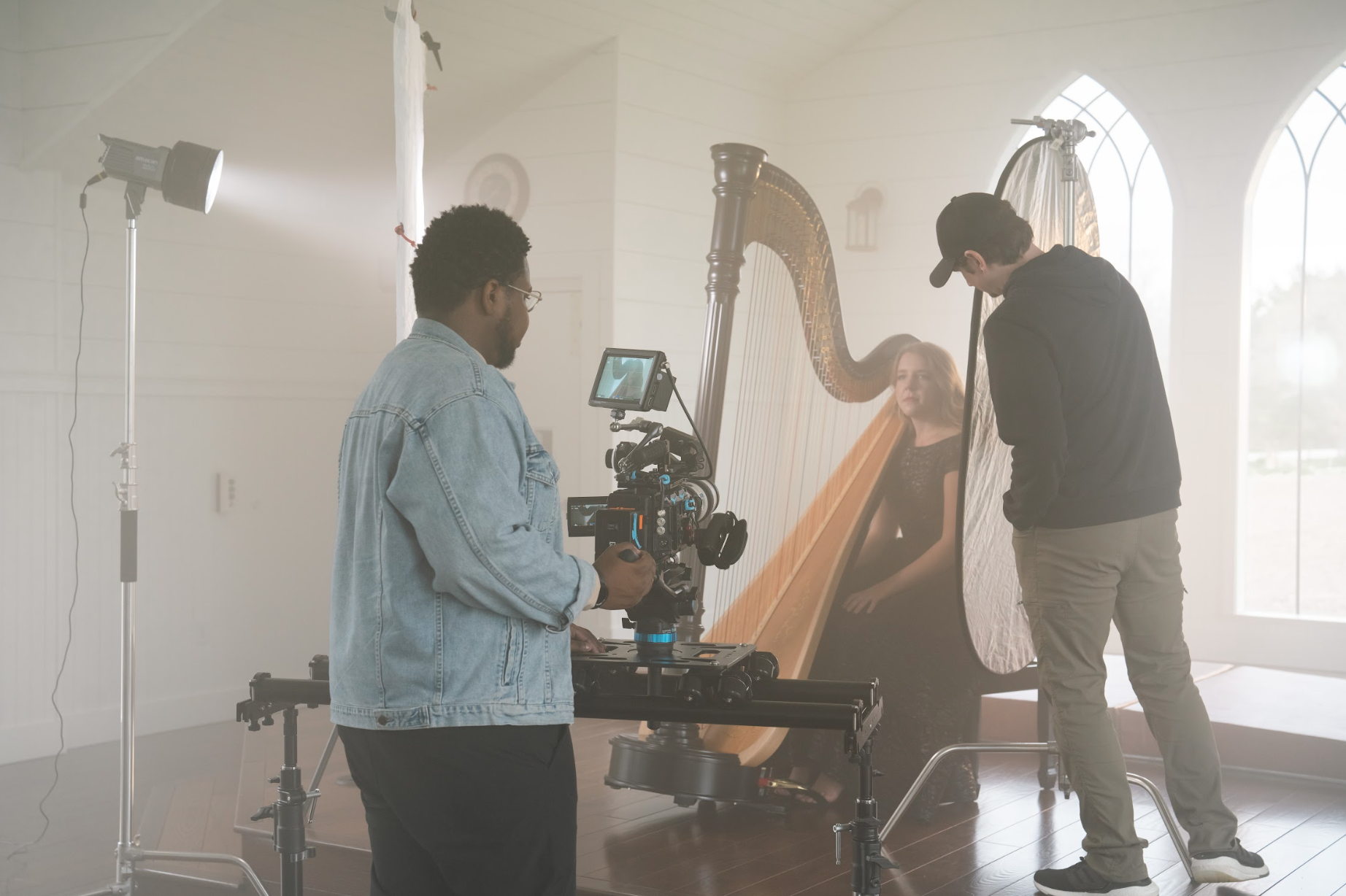 People filming a woman playing the harp in a studio setting with lighting equipment and a camera on a dolly.