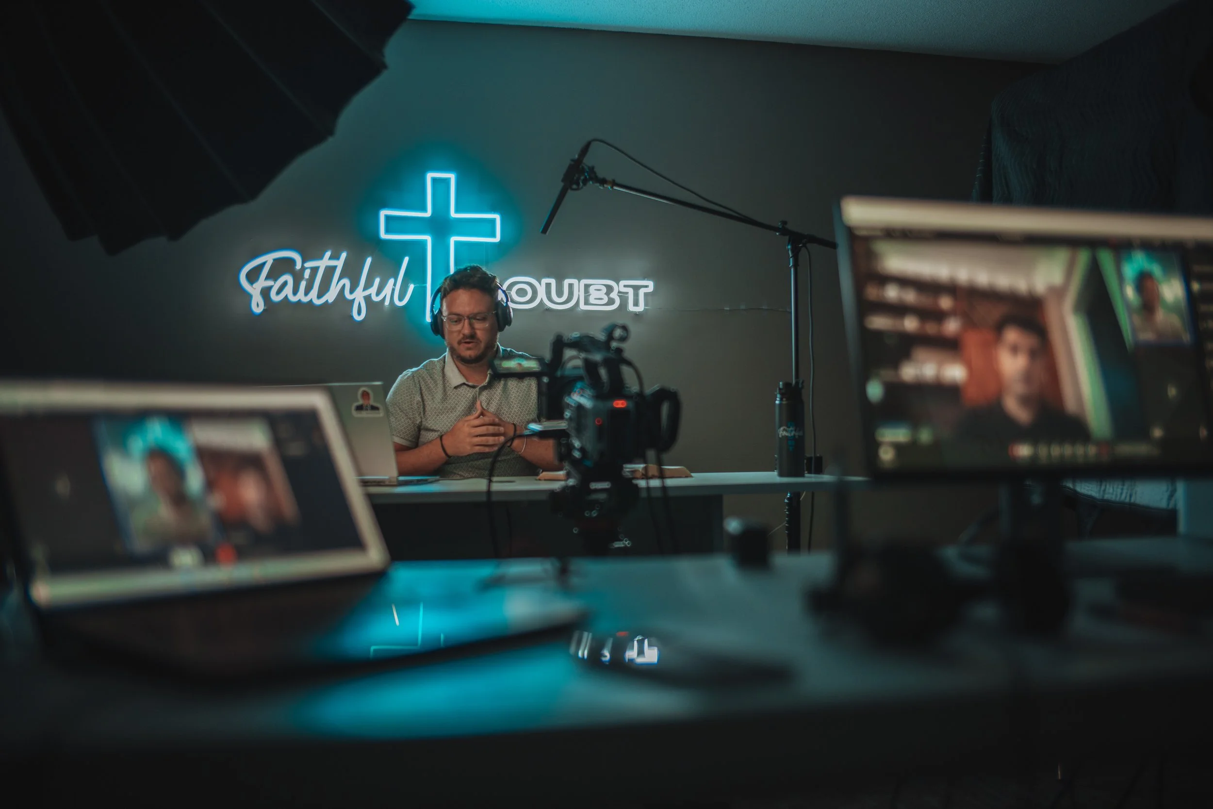 Man wearing headphones hosting a podcast or video stream in a studio with neon sign reading 'Faithful Doubt' and cross symbol. Multiple screens and camera equipment visible in the foreground.