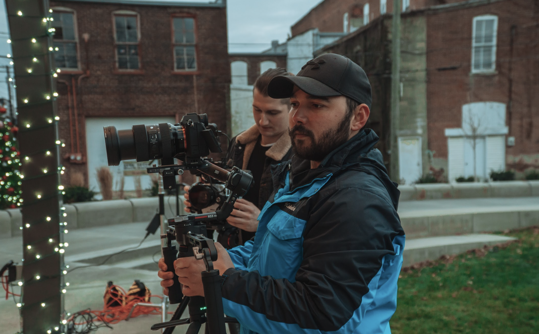 Two people filming outdoors with cameras on stabilizers, wearing jackets, one appears to be focused on the equipment, brick building and holiday lights in the background.