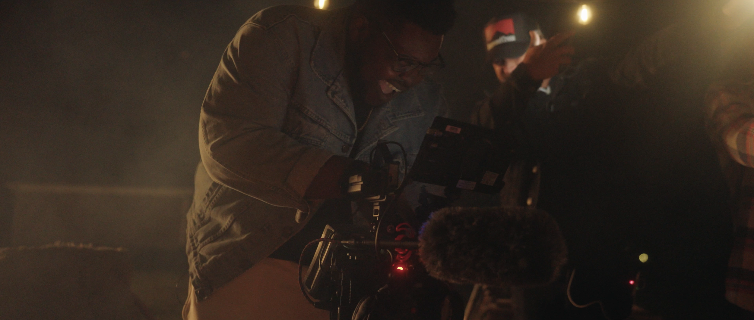 Josh Bennett in a denim jacket is smiling while looking at a camera monitor, with Brandon Froedge in the background holding a phone with a bright light. It looks like a filming setup in a dim environment.