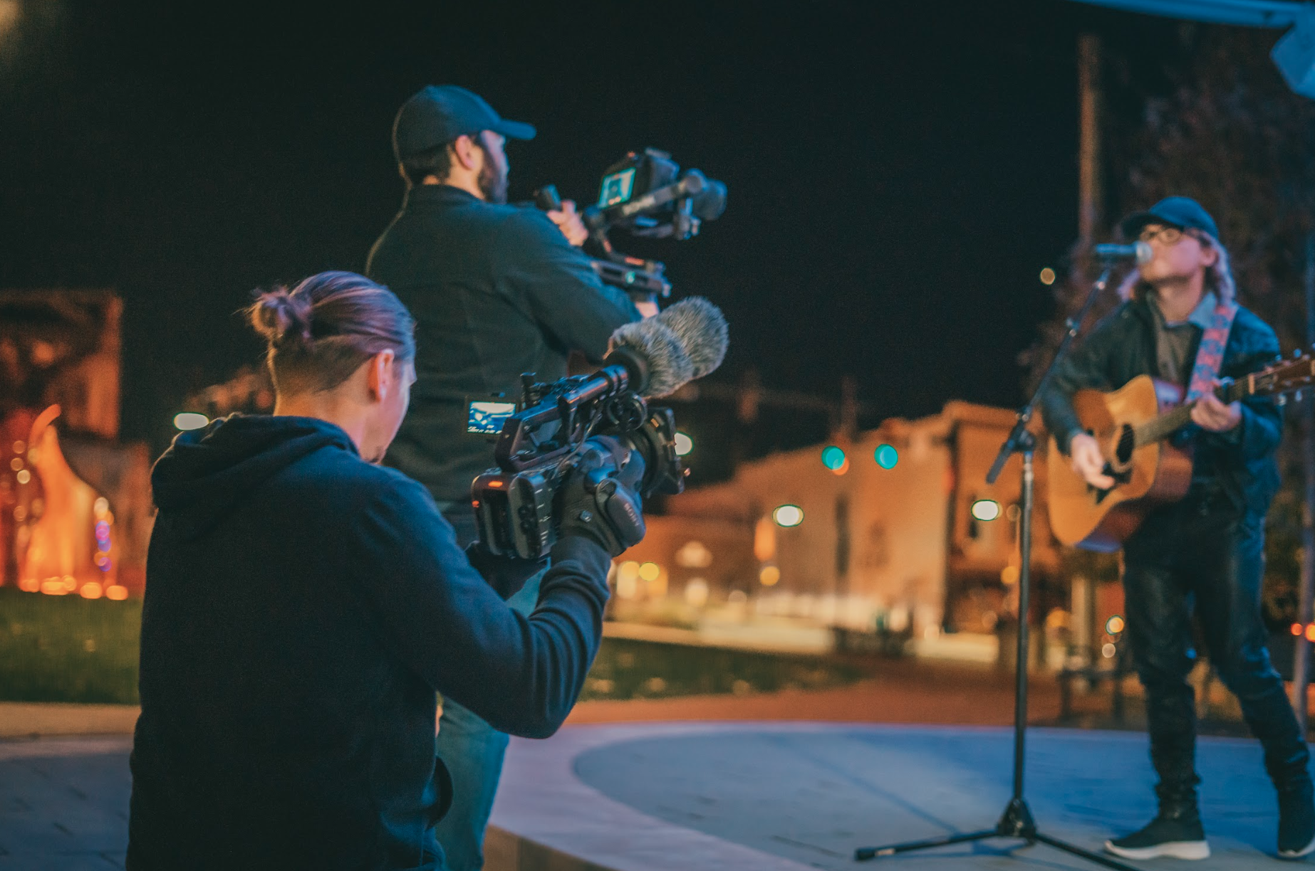 Two people filming a musician playing guitar and singing on stage at night.