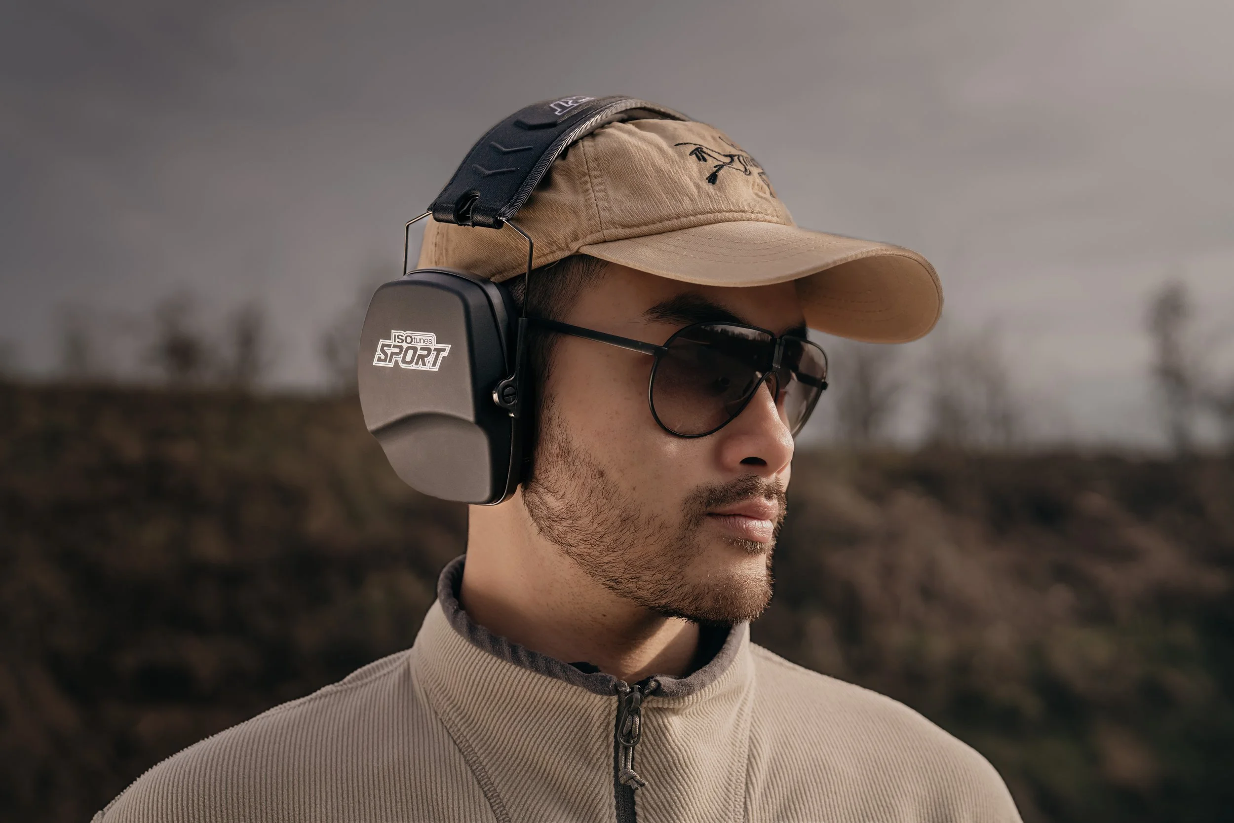 A man wearing a beige baseball cap, dark sunglasses, and black noise-canceling earmuffs with the label "ISOtunes SPORT," standing outdoors.
