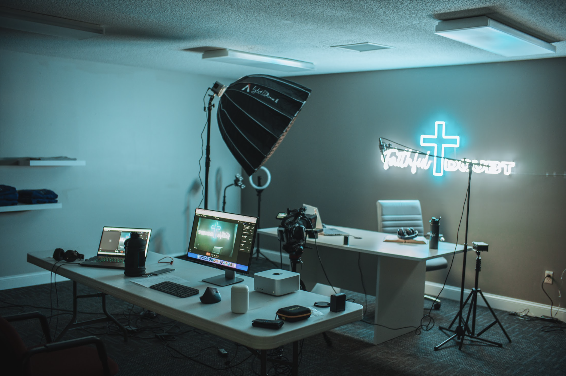 Modern podcast studio setup with computers, microphones, cameras, and lighting equipment. A neon sign with a cross and text is on the wall.