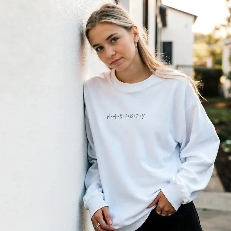 Young woman with blonde hair wearing a white sweatshirt with the word 'HABIBTY' on it, standing outdoors against a white wall.