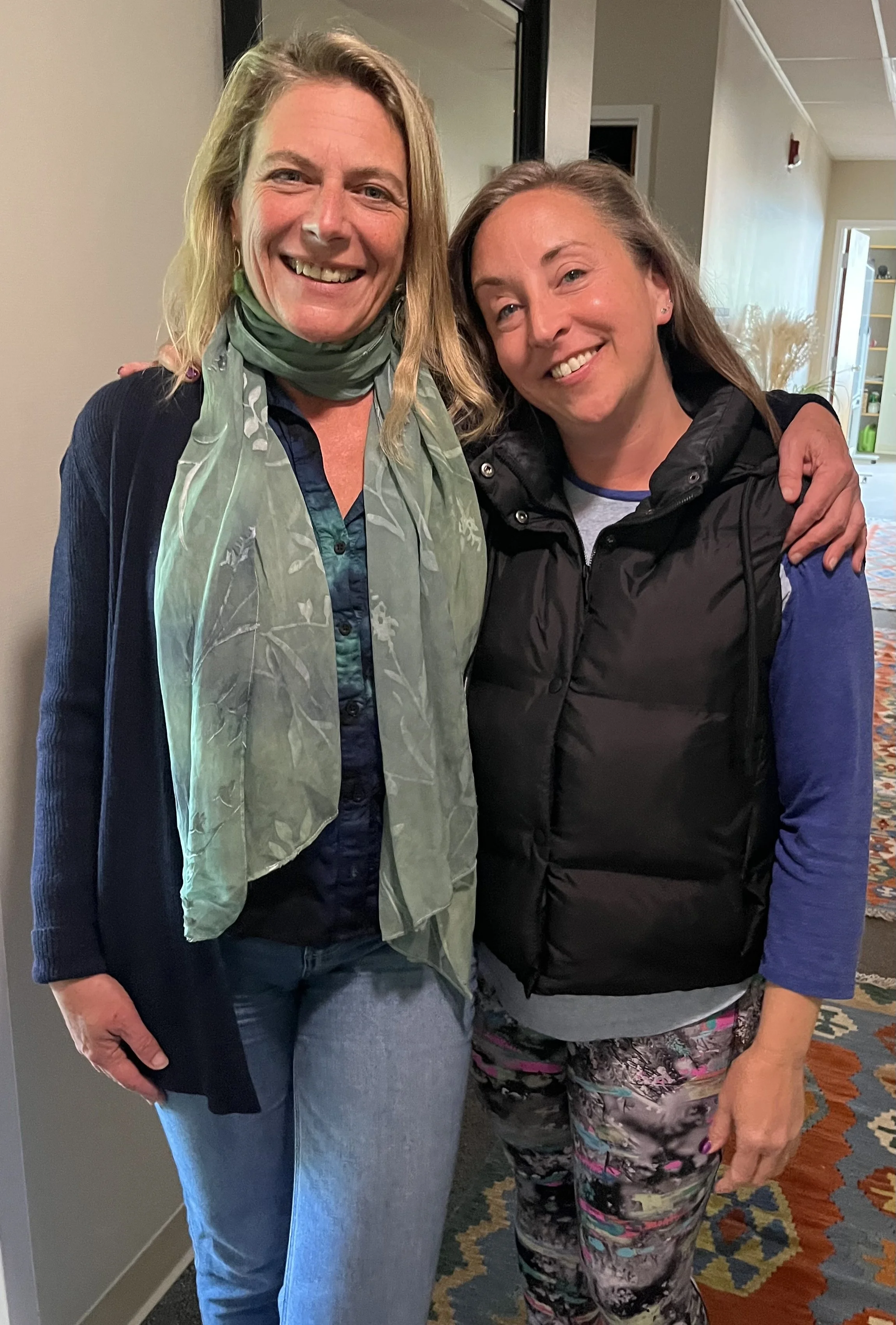 Two women smiling and posing closely indoors, one with blonde hair wearing a green scarf and blue blazer, the other with brown hair wearing a black puffy vest and patterned pants.