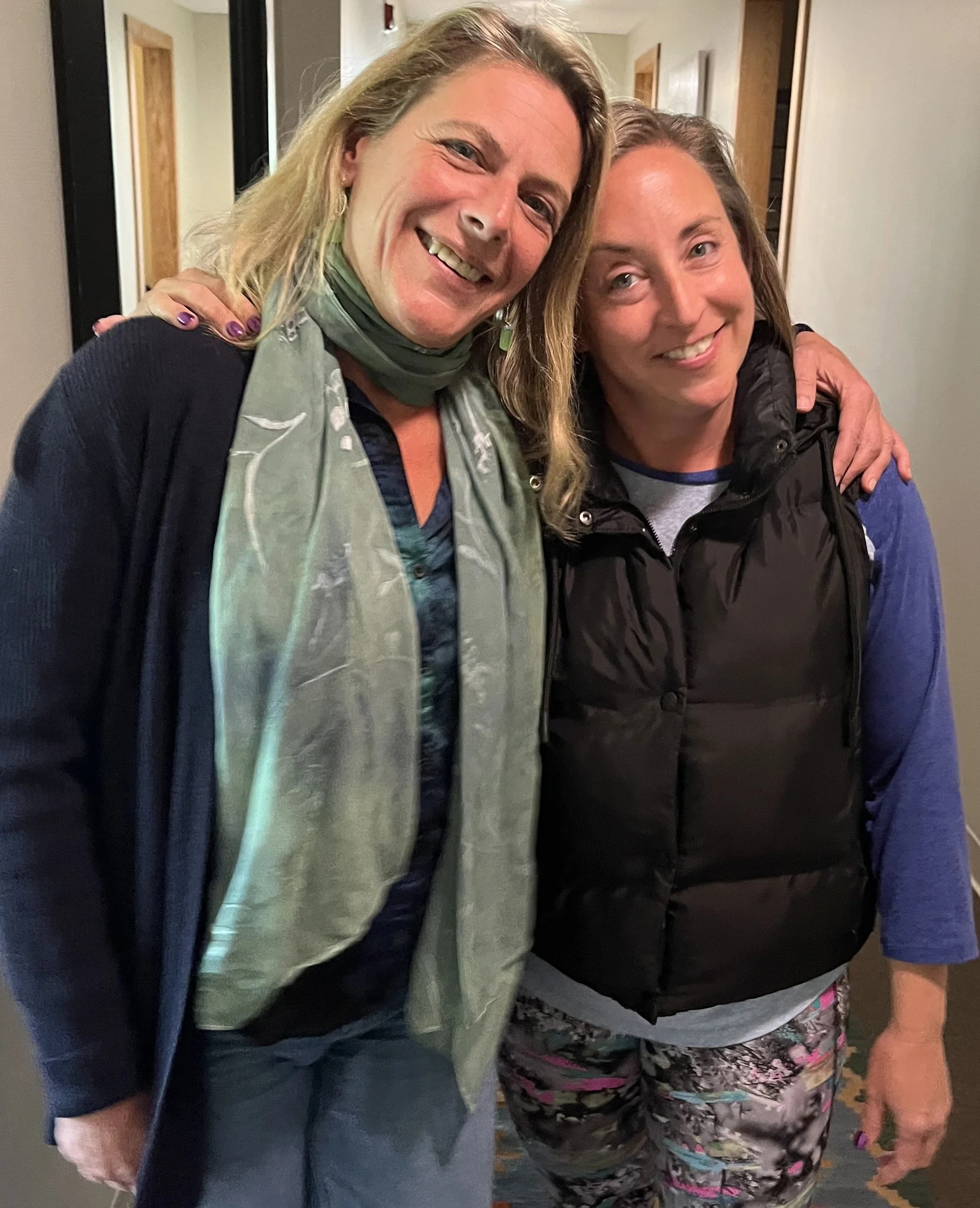 Two women smiling and hugging each other, standing close in an indoor setting.