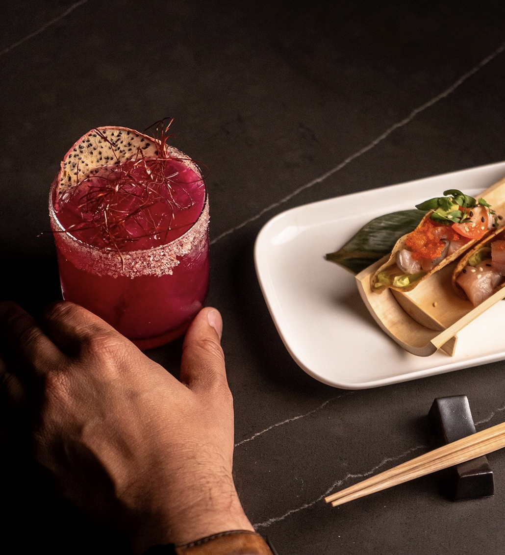Guest holding a pink dragonfruit cocktail beside a plated dish.