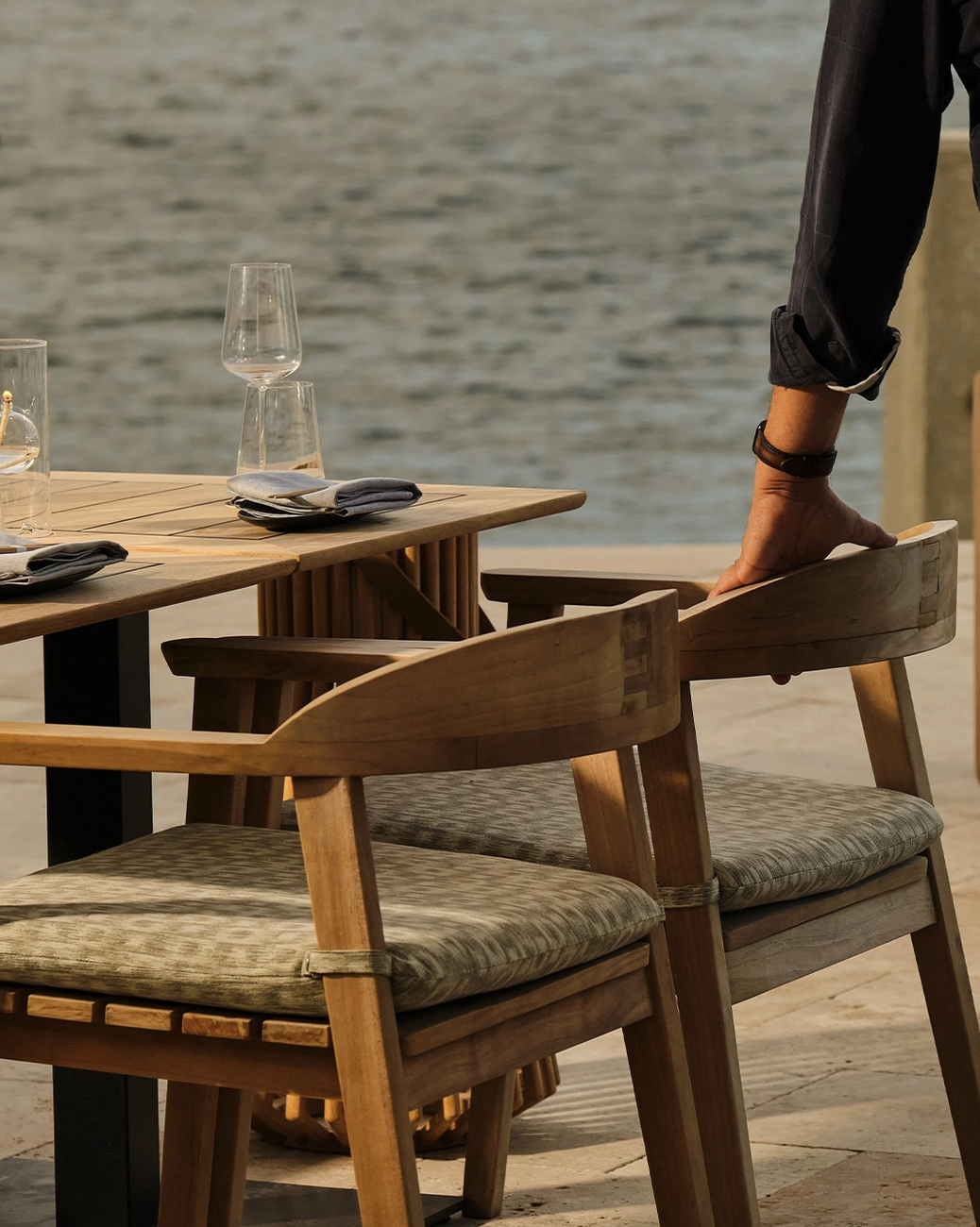 Waterfront dining table set with glassware and wooden chairs as a server adjusts a seat.