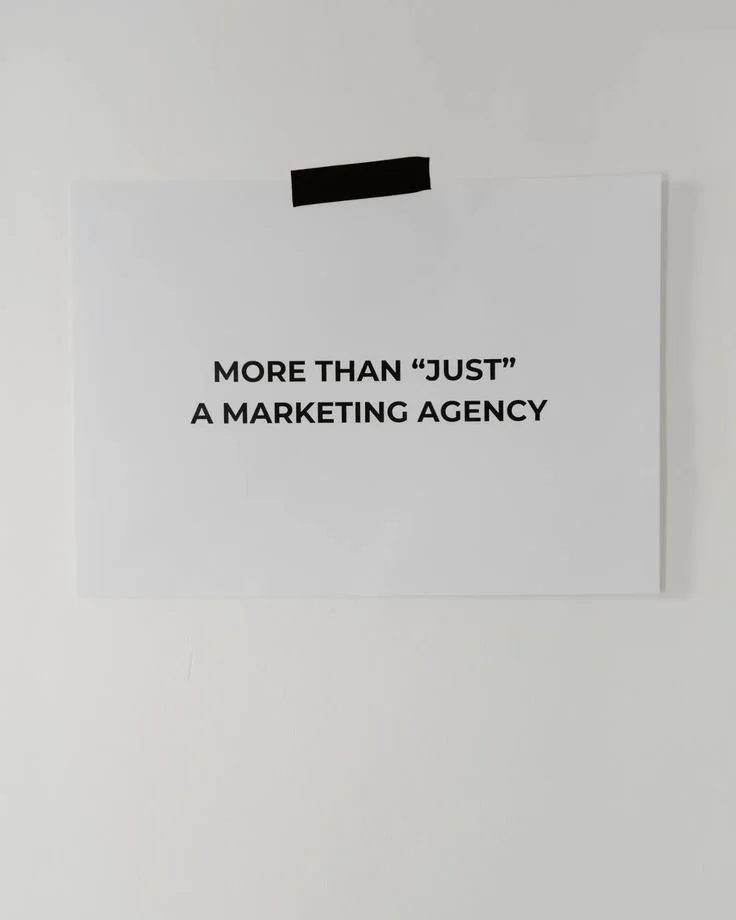 Sign taped to wall that reads 'MORE THAN 'JUST' A MARKETING AGENCY' in black text.