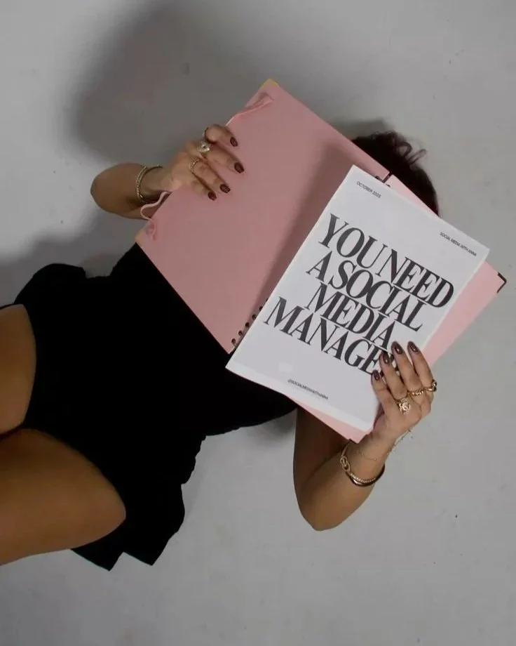 Person lying on the floor holding a pink folder and a printed page that reads "You need a social media manager."