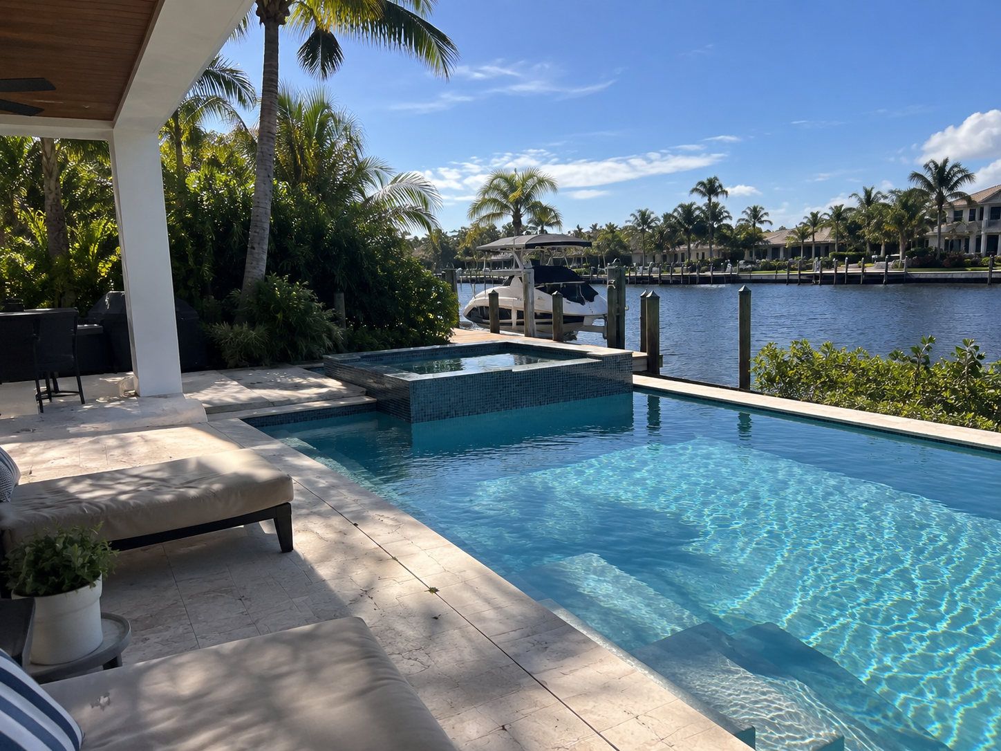 A backyard swimming pool with a hot tub, lounge chairs, and lush tropical plants near a canal with a boat docked.