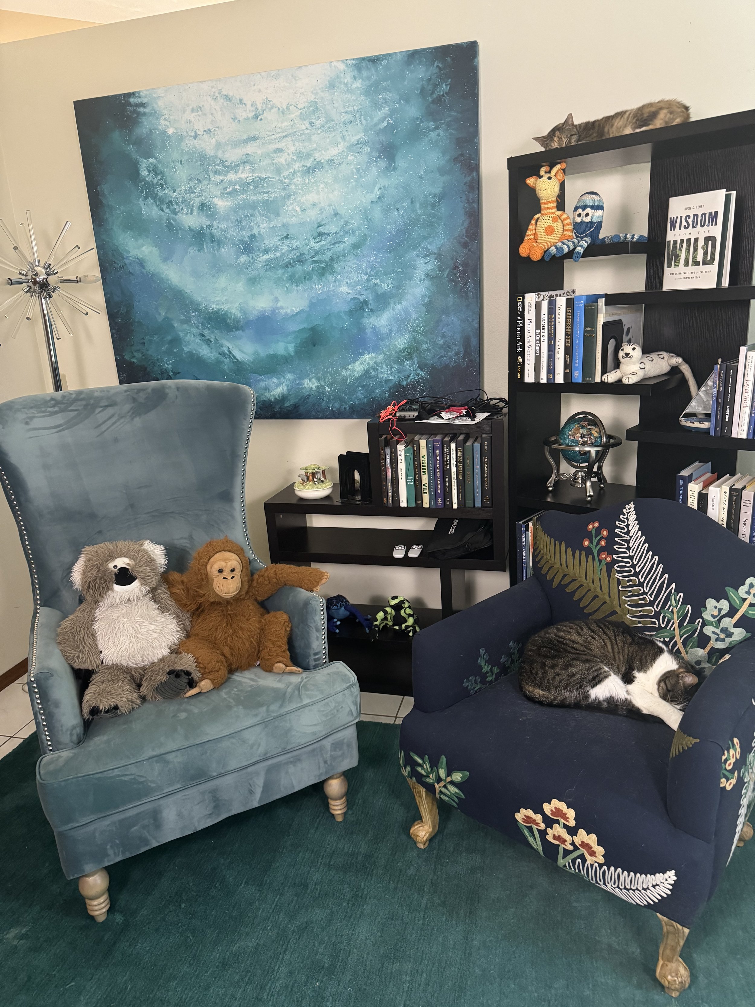 A cozy living room corner with two plush chairs, a large abstract blue and white painting on the wall, and a black bookshelf filled with books and decorative plush toys. A cat is sleeping on top of the bookshelf, while a gray and white cat is curled up on a floral-patterned dark blue armchair.