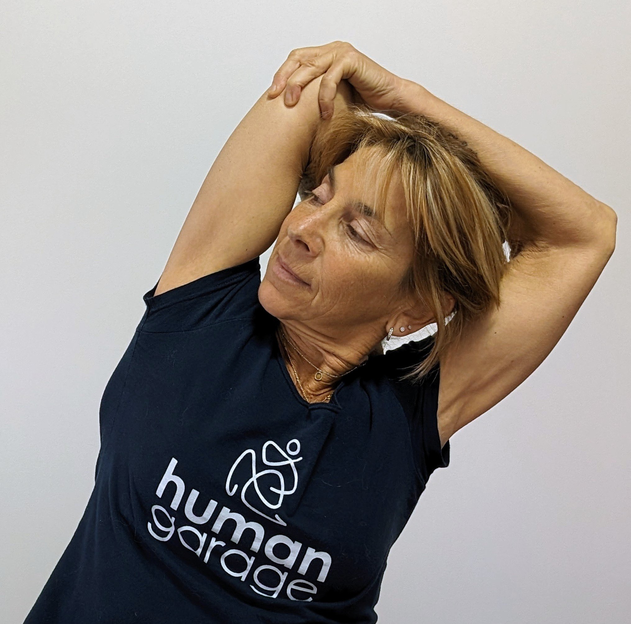 Woman stretching with hands clasped behind her head, wearing a navy blue t-shirt that says 'human guidance'.