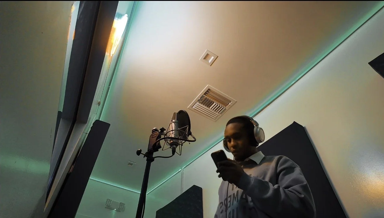 A person in a recording studio wearing headphones, looking at their phone, with a microphone on a stand nearby and soundproofing panels on the walls.