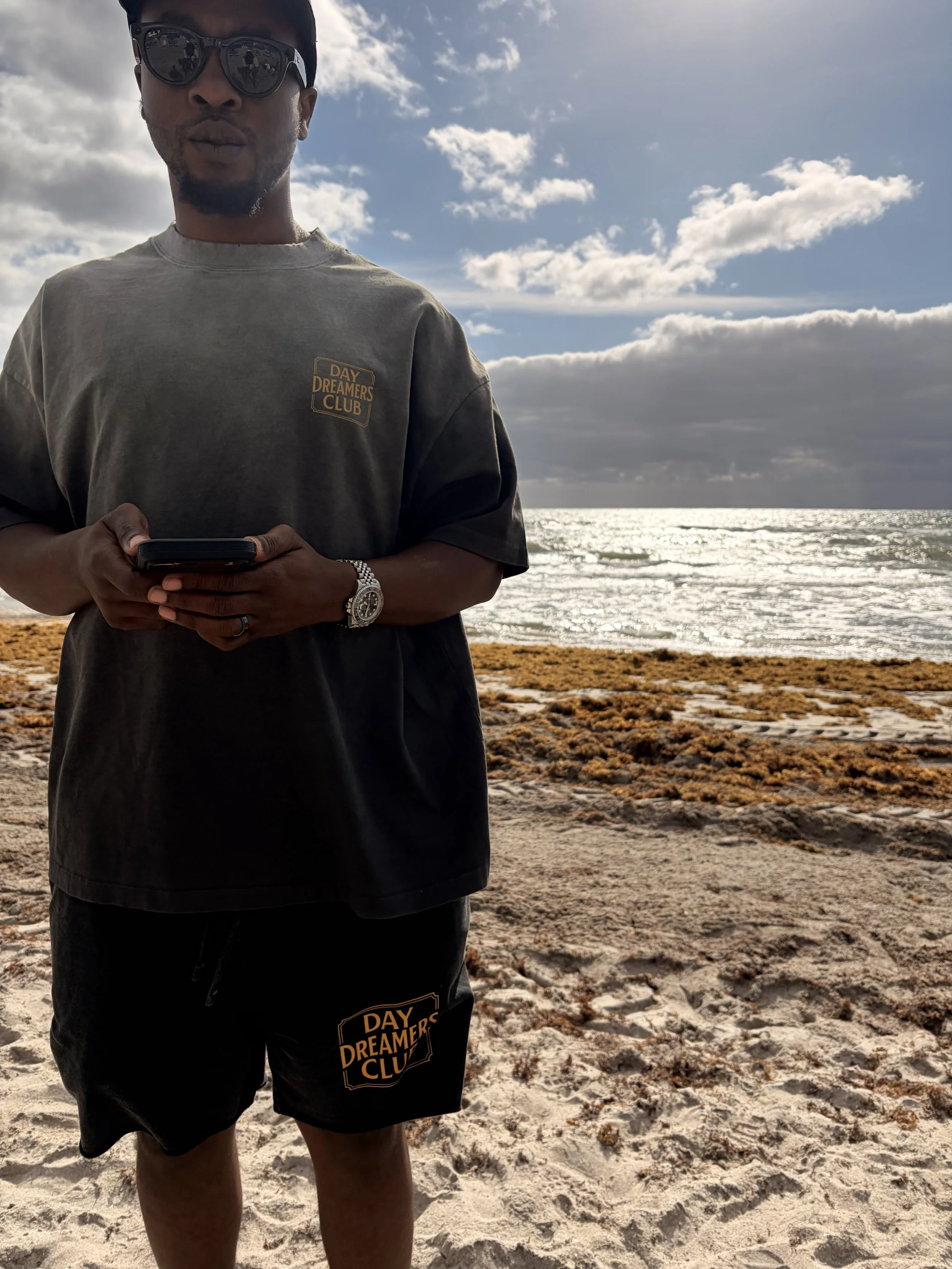 A man standing on a sandy beach facing the camera, holding a phone, wearing sunglasses, a gray t-shirt and black shorts with a 'Day Dreamers Club' logo, with the ocean and cloudy sky in the background.