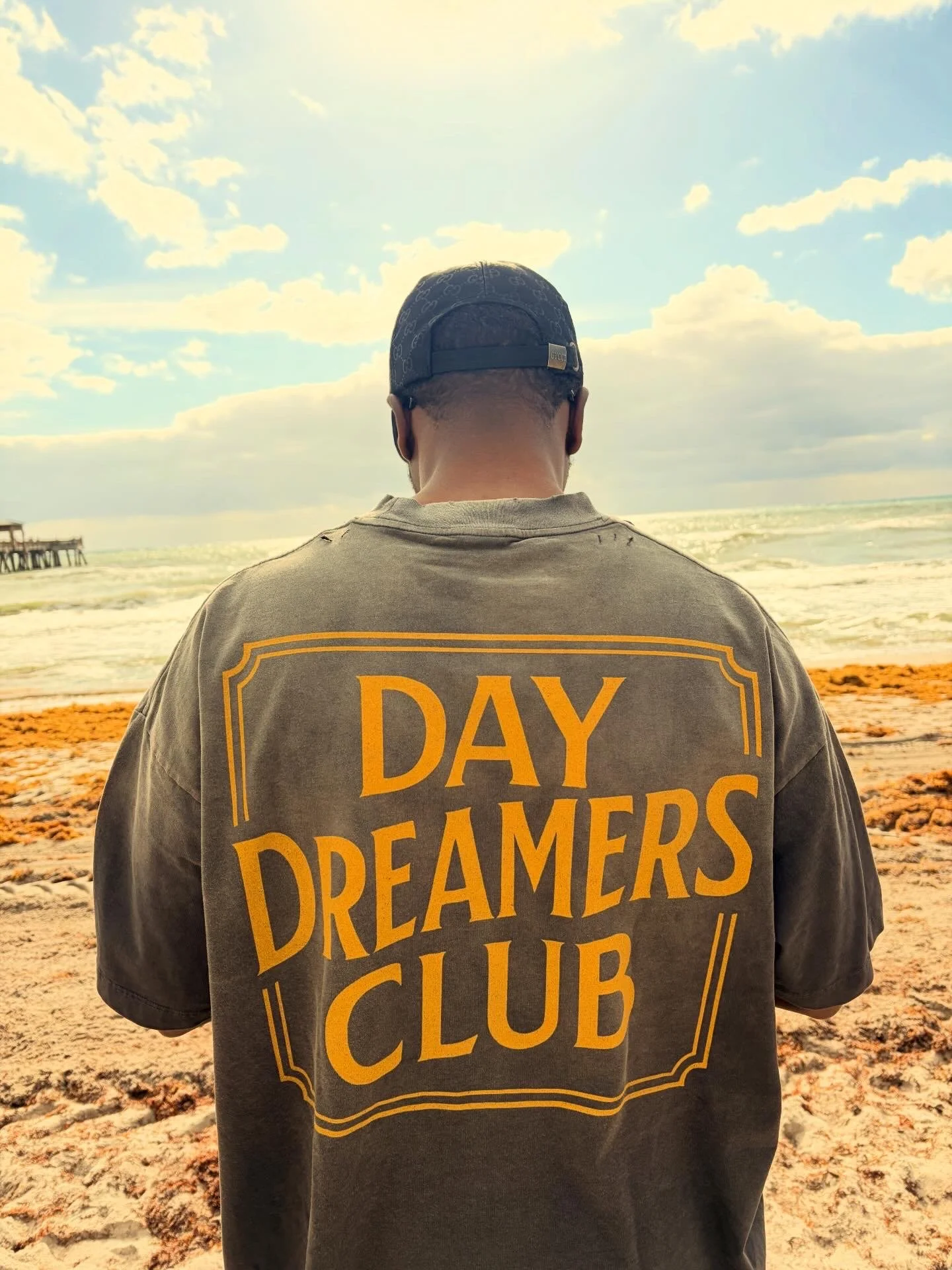 Person wearing a cap and a t-shirt that says 'Day Dreamers Club' on a beach with the ocean and sky in the background.