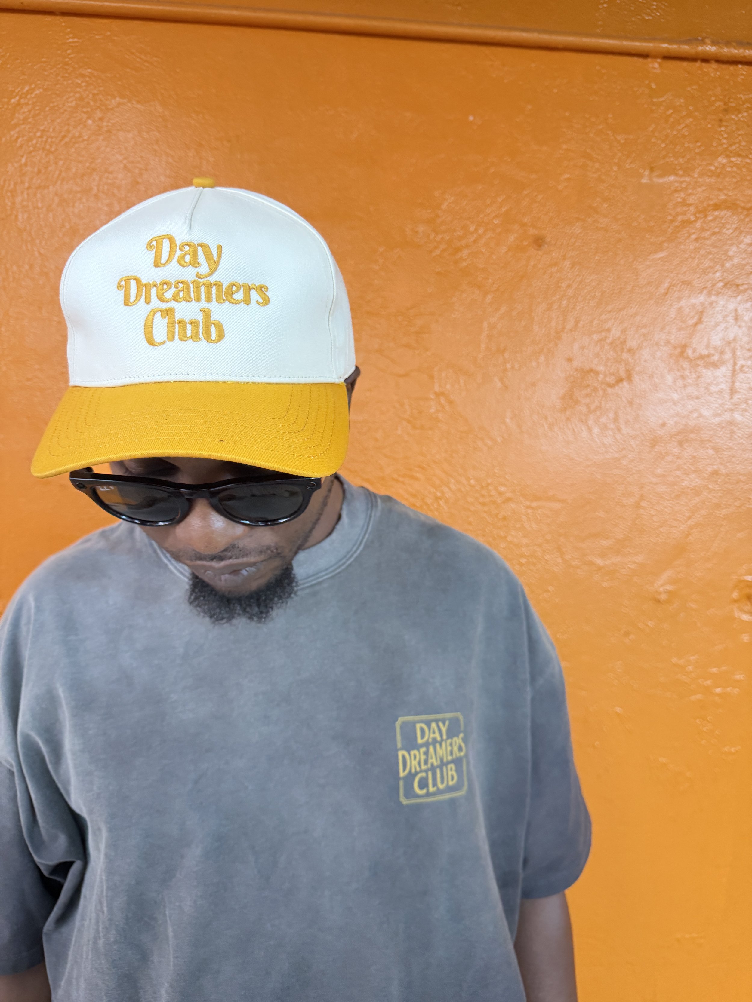 Person wearing sunglasses, a gray sweatshirt with 'Day Dreamers Club' logo, and a yellow and white cap with 'Day Dreamers Club' embroidery, standing against an orange wall.