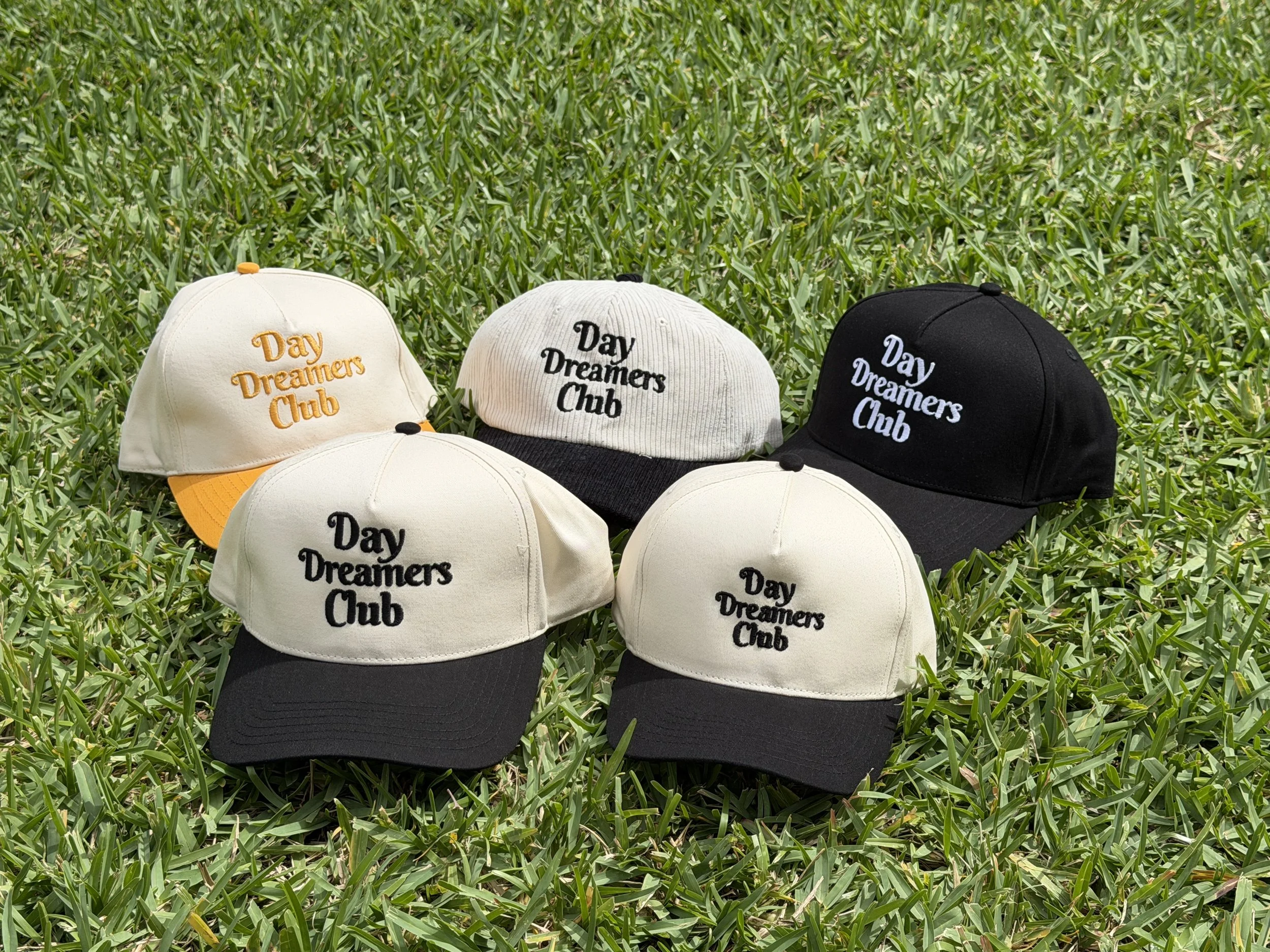 Six caps with embroidered text 'Day Dreamers Club', in various colors, placed on green grass.