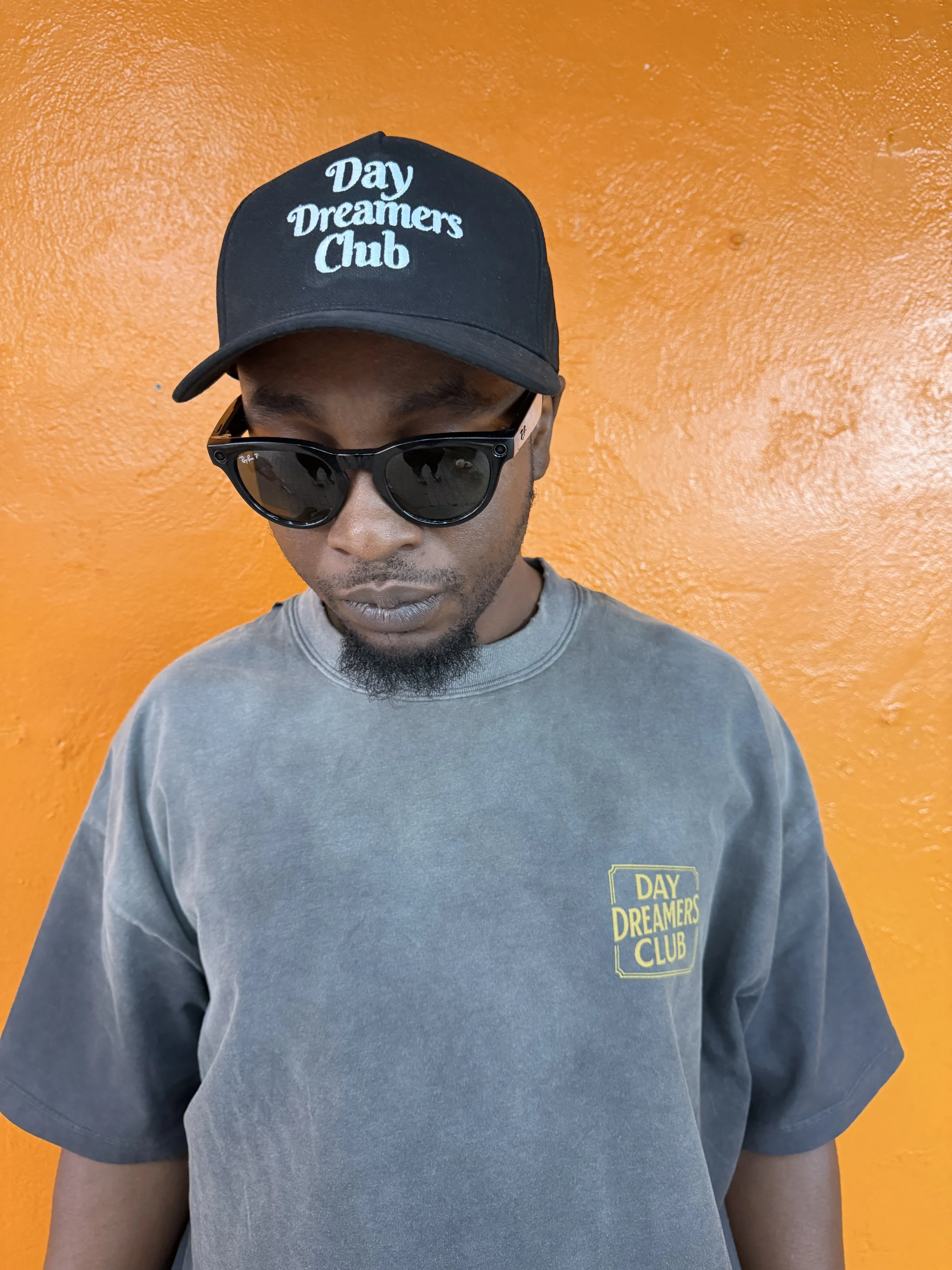 A man wearing a black cap with the words 'Day Dreamers Club' and sunglasses, standing against an orange wall.