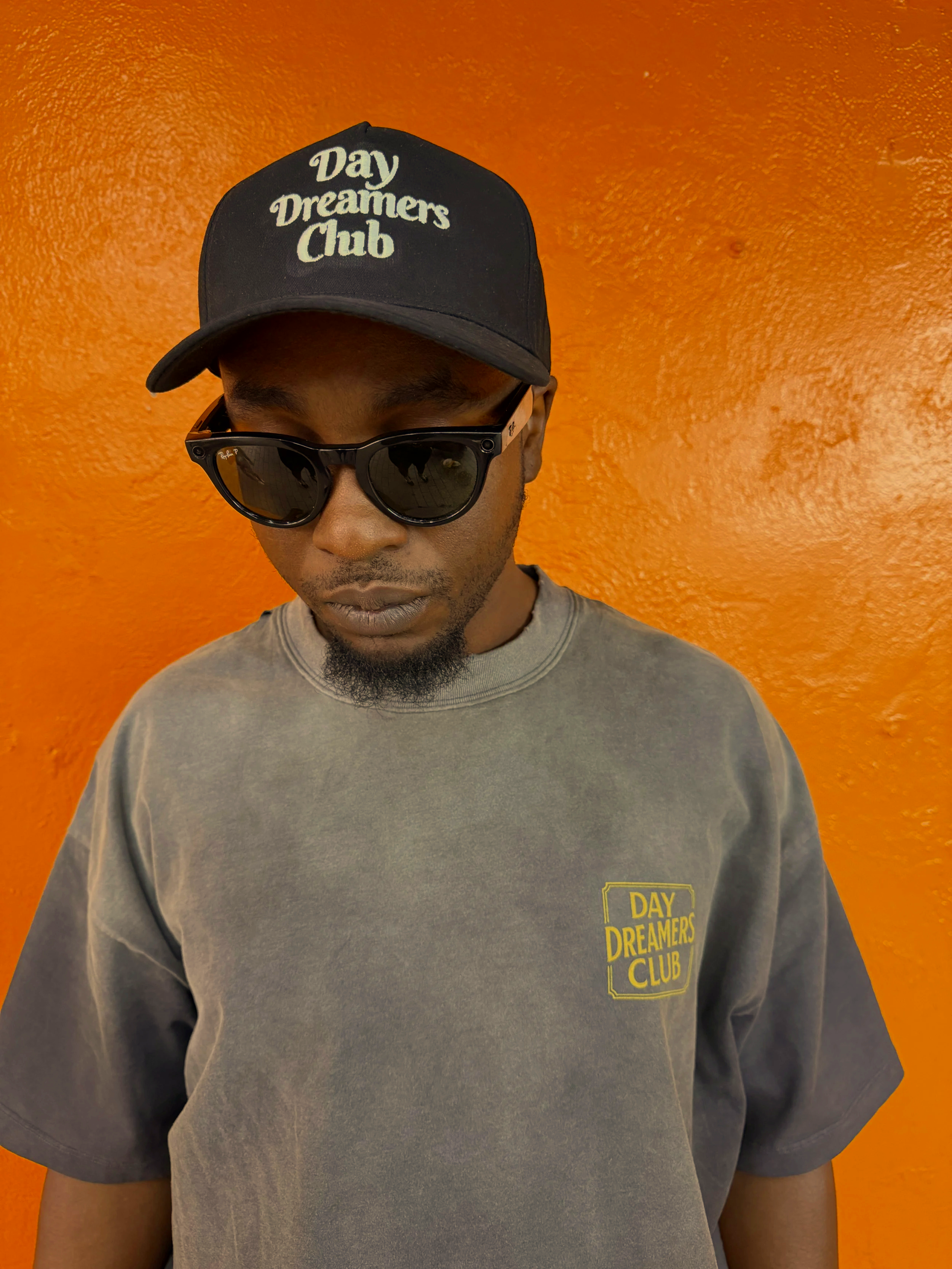 A man wearing black sunglasses and a gray t-shirt with yellow text that says "Day Dreamers Club" stands in front of an orange wall. He is also wearing a black cap with white text that says "Day Dreamers Club."