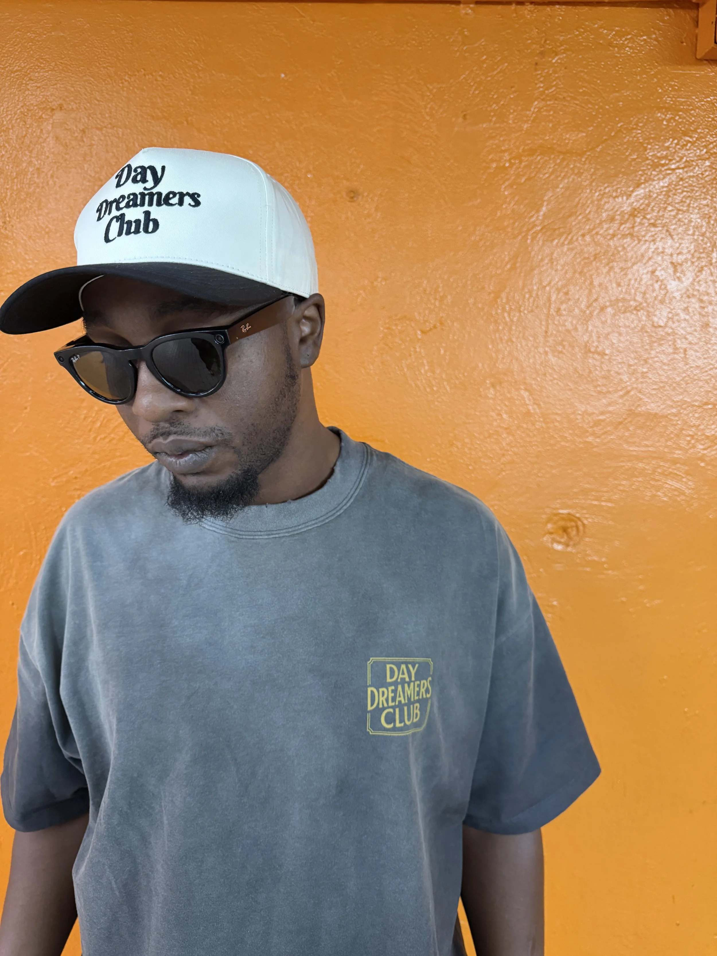 A man wearing a gray T-shirt and black sunglasses, with a white and black cap that says "Day Dreamers Club," standing against an orange wall.