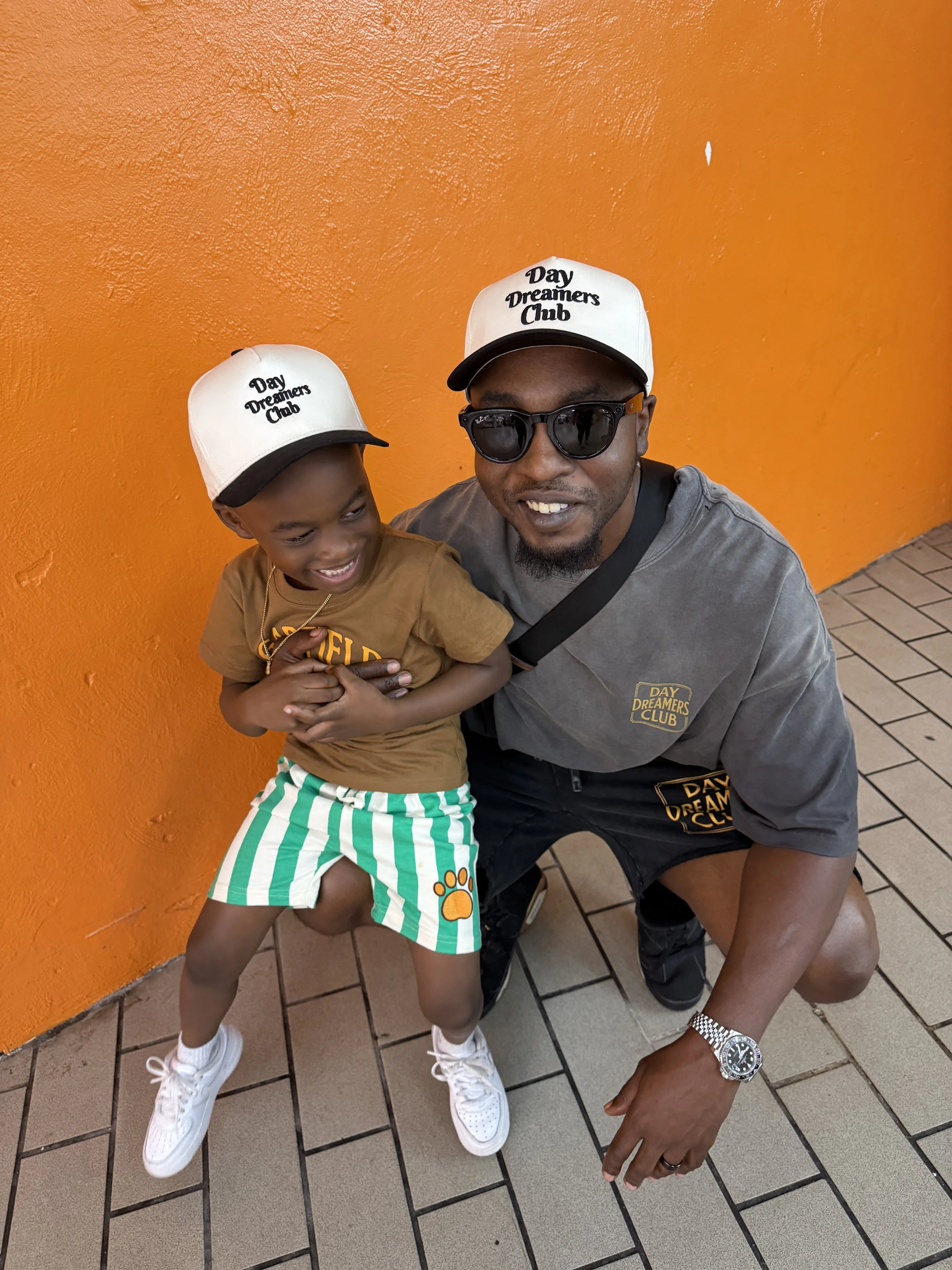 A man and a young boy posing together against an orange wall, both wearing white hats that say 'Day Dreamers Club.' The boy is smiling and wearing striped green and white shorts with a paw print. The man is wearing sunglasses, a gray shirt, and a watch, and is kneeling beside the boy.