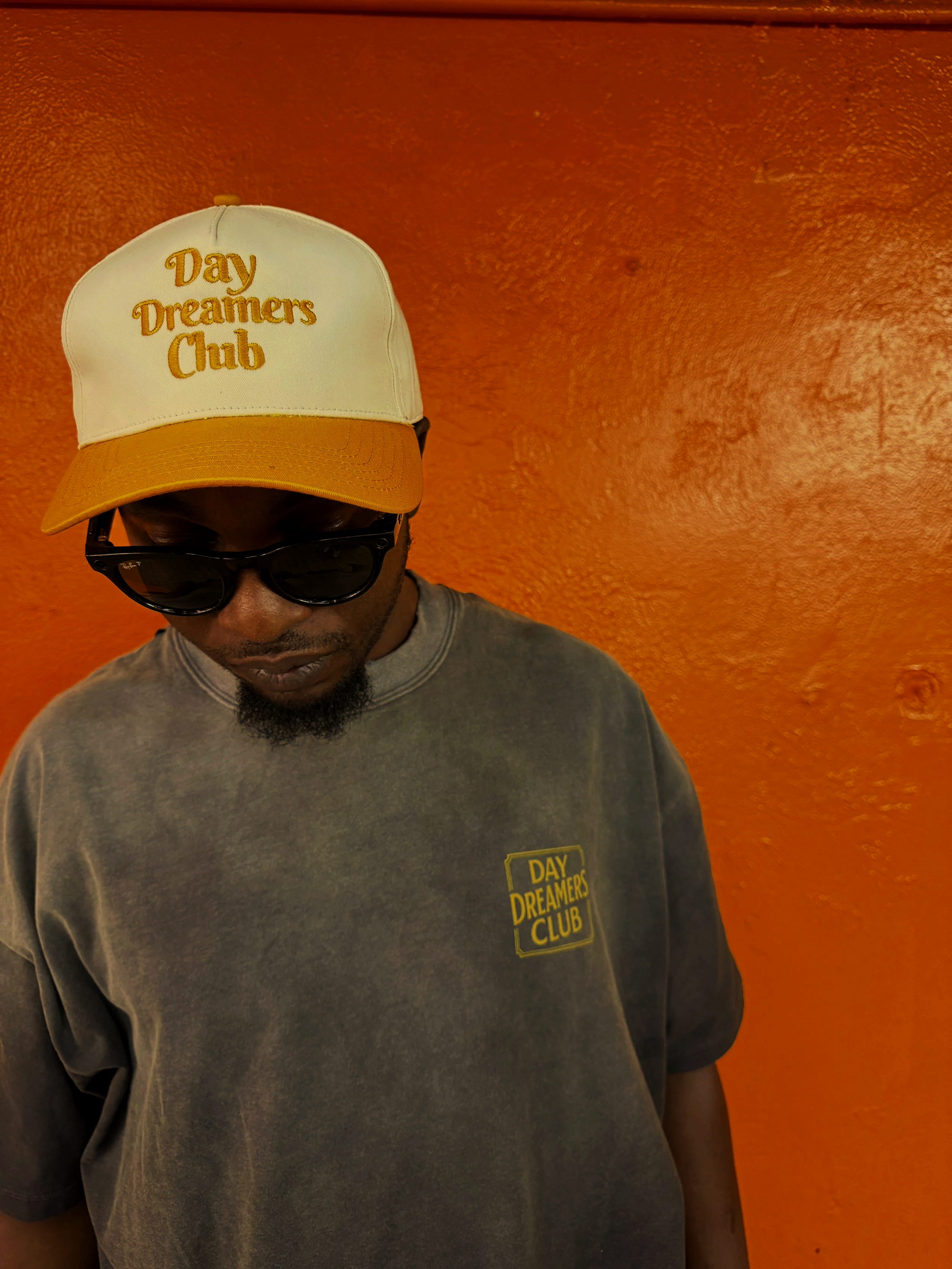A man wearing a white and yellow 'Day Dreamers Club' cap, black sunglasses, and a gray sweatshirt with a yellow 'Day Dreamers Club' logo, standing against an orange textured wall.
