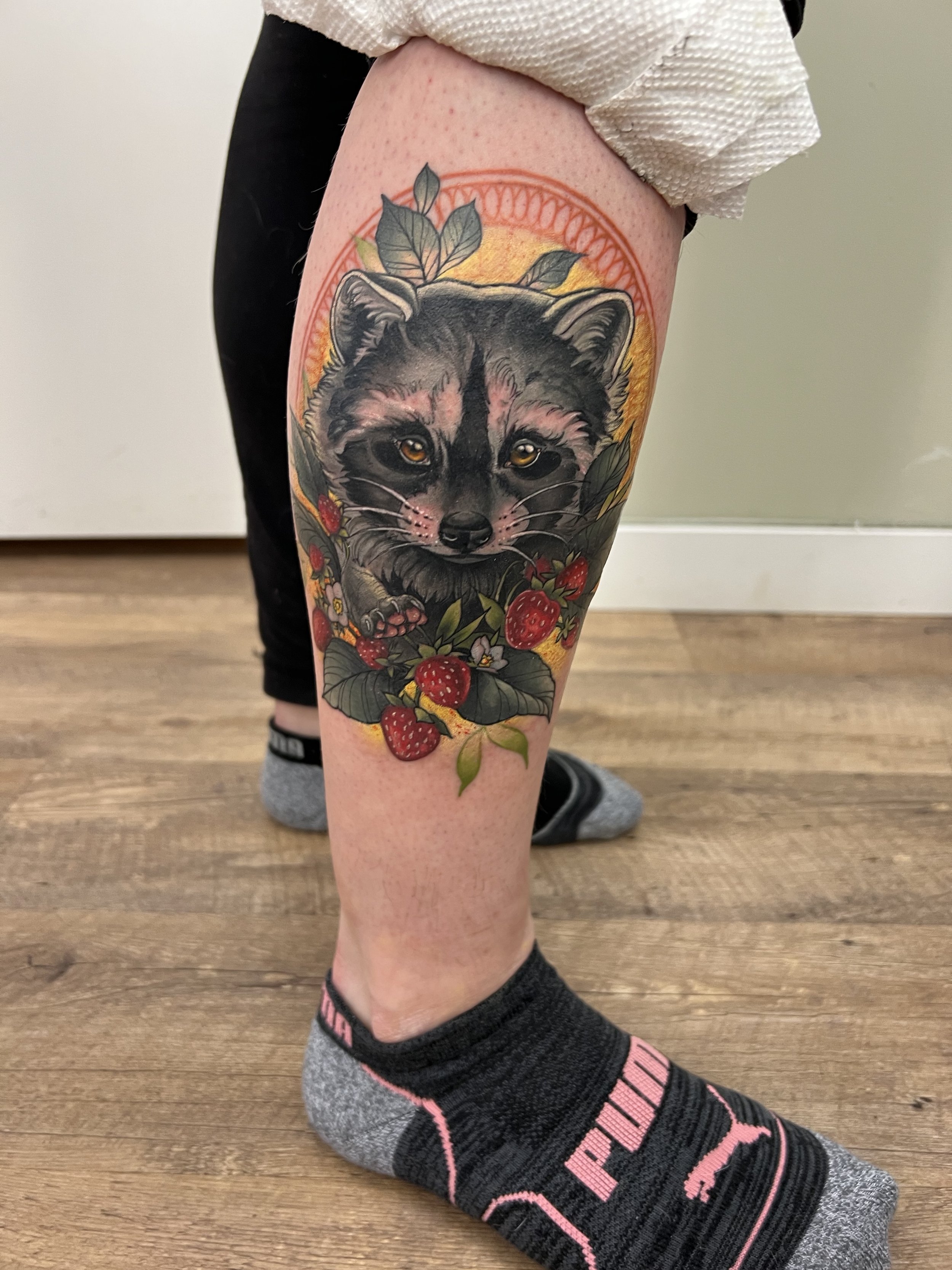 Tattoo of a raccoon surrounded by strawberries on a person's leg