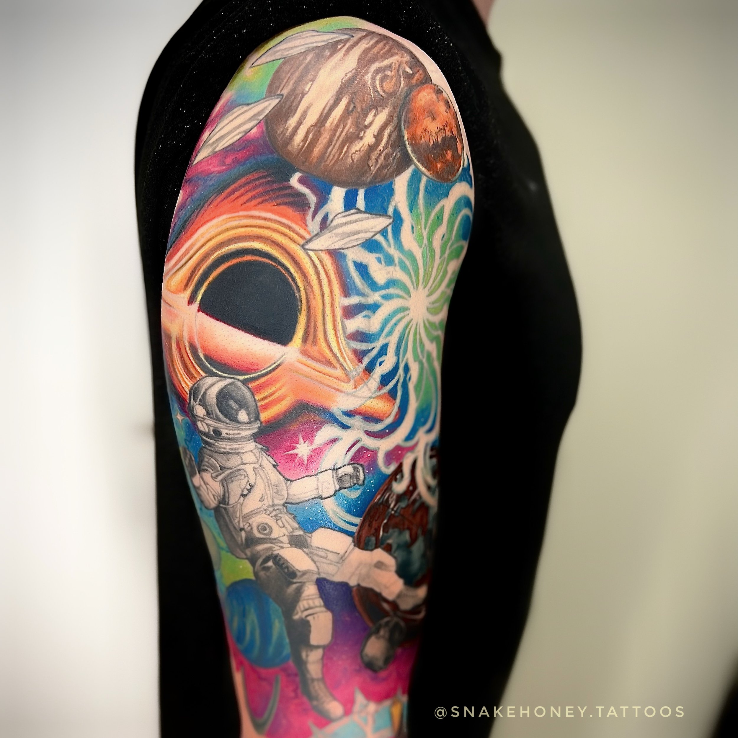 Colorful space-themed tattoo on arm featuring an astronaut, planets, stars, and a black hole.